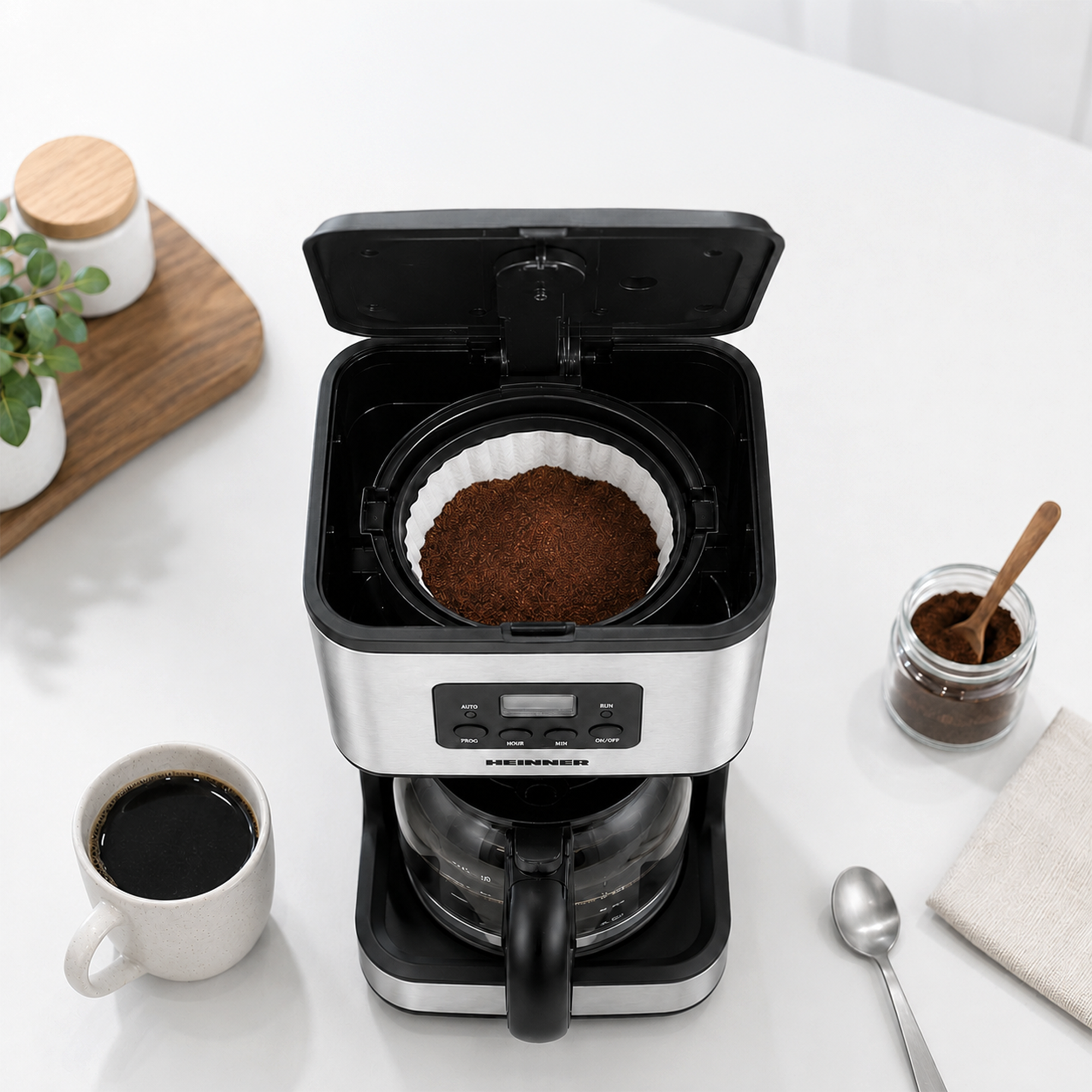 An open coffee maker with ground coffee in the filter stands on a white countertop next to a cup of coffee.