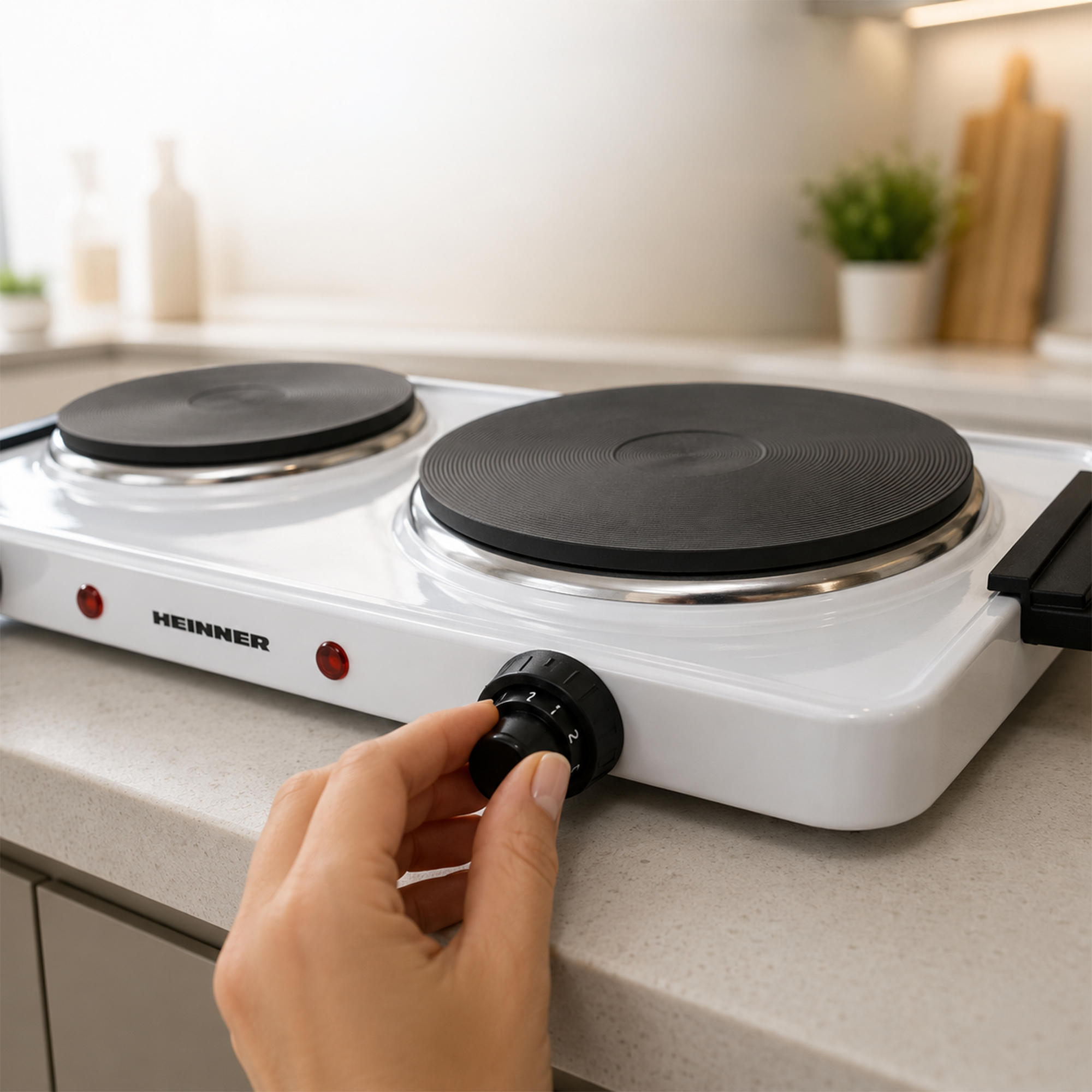 A hand turns the black control knob of a white Heinner double hotplate on a kitchen counter.