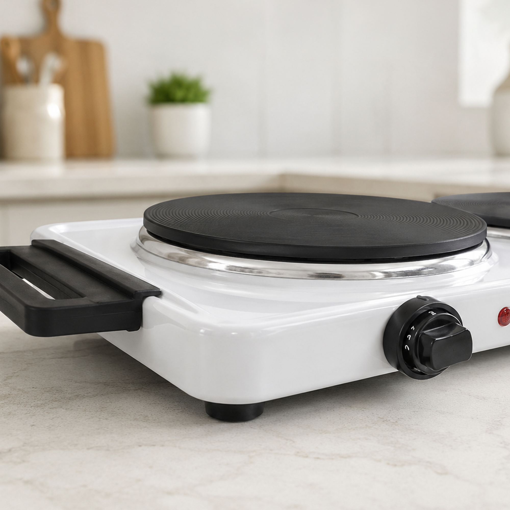 A white portable double hot plate with black burners and a control knob sits on a light countertop in a blurred kitchen.
