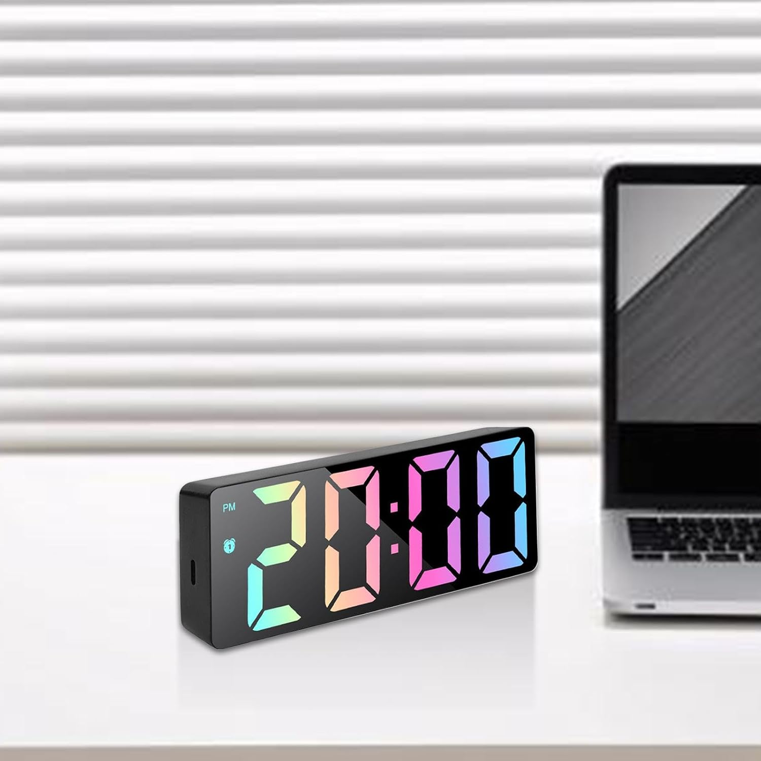 Digitale LED-Uhr mit bunten Ziffern auf weißem Schreibtisch, neben einem Laptop vor Jalousien.