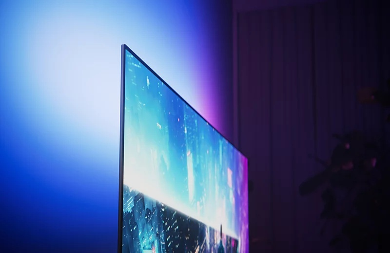 Smukły telewizor wyświetla miejski pejzaż, rzucając niebieskie światło otoczenia na ścianę.