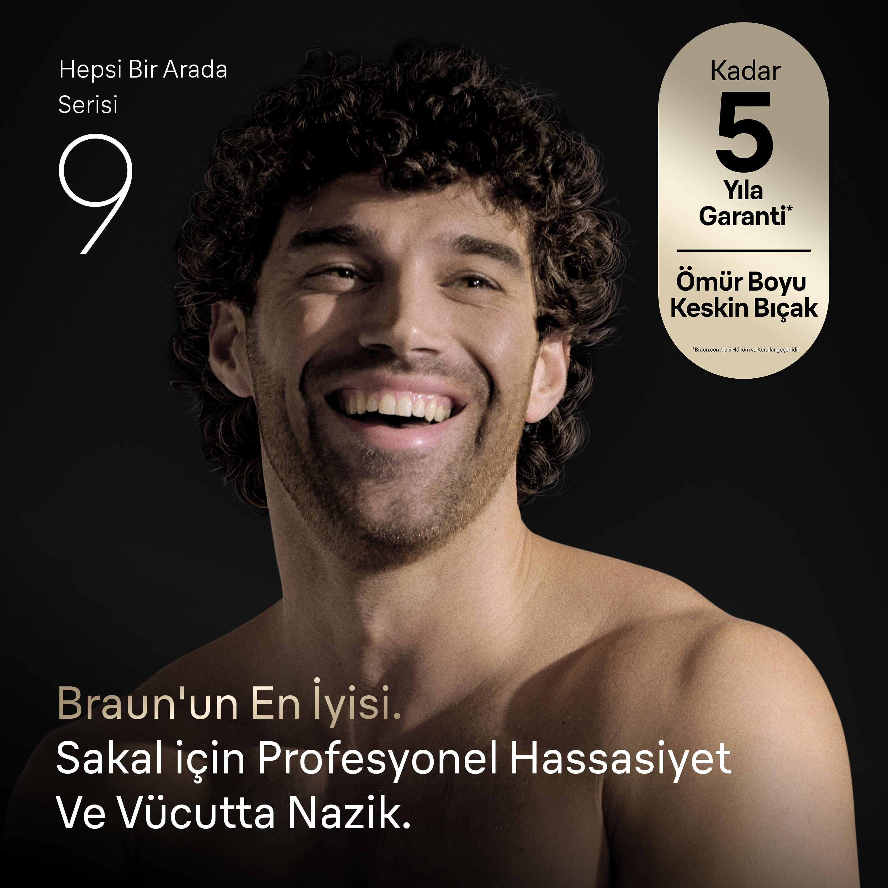 Kıvırcık saçlı ve sakallı, üstü çıplak gülümseyen bir erkek, koyu arka plan önünde.