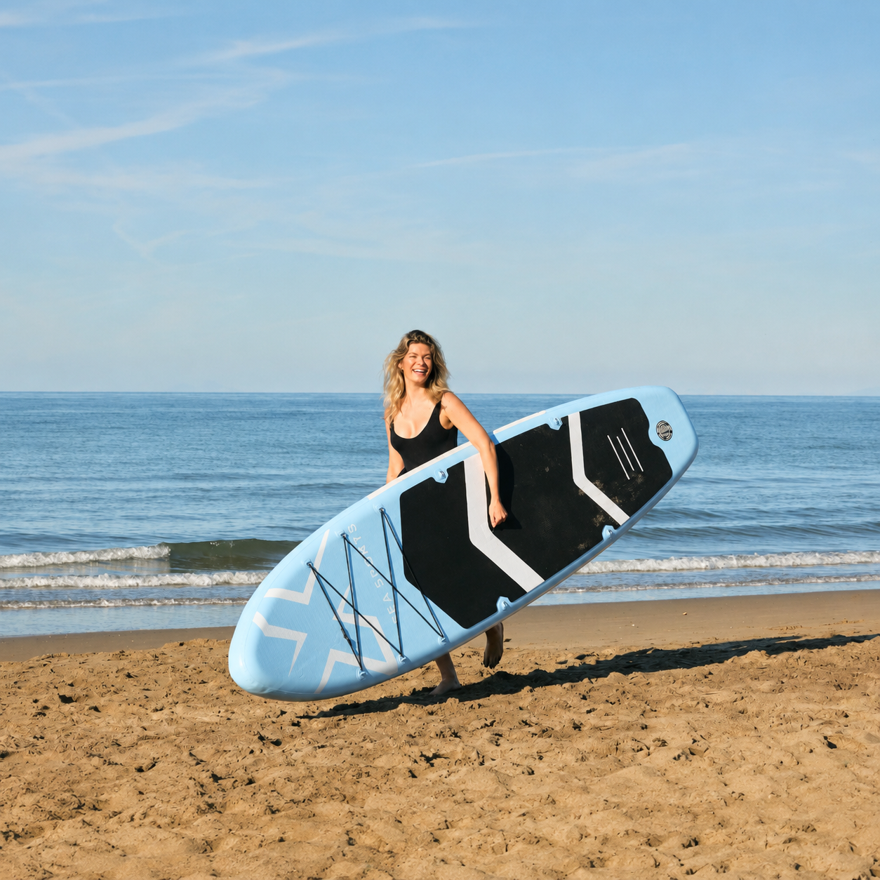 Una mujer sonriente con traje de baño negro lleva una tabla de paddle surf azul claro y negra en una playa.