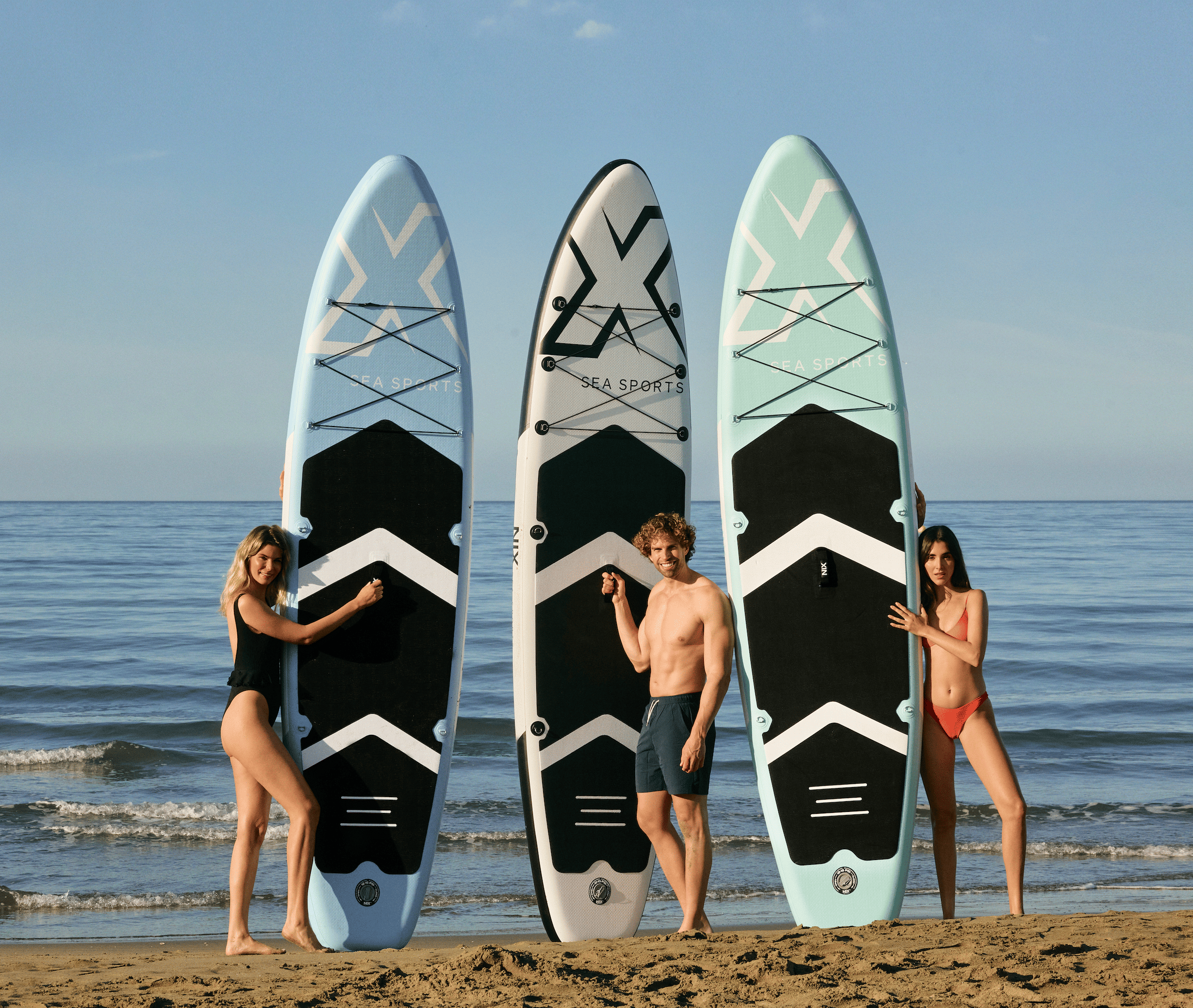 Tres personas en ropa de baño de pie en una playa, cada una sosteniendo una tabla de surf de remo.