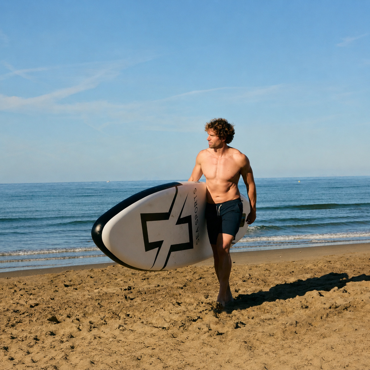 Un hombre con cabello rizado camina por una playa de arena, cargando una tabla de surf blanca y negra.