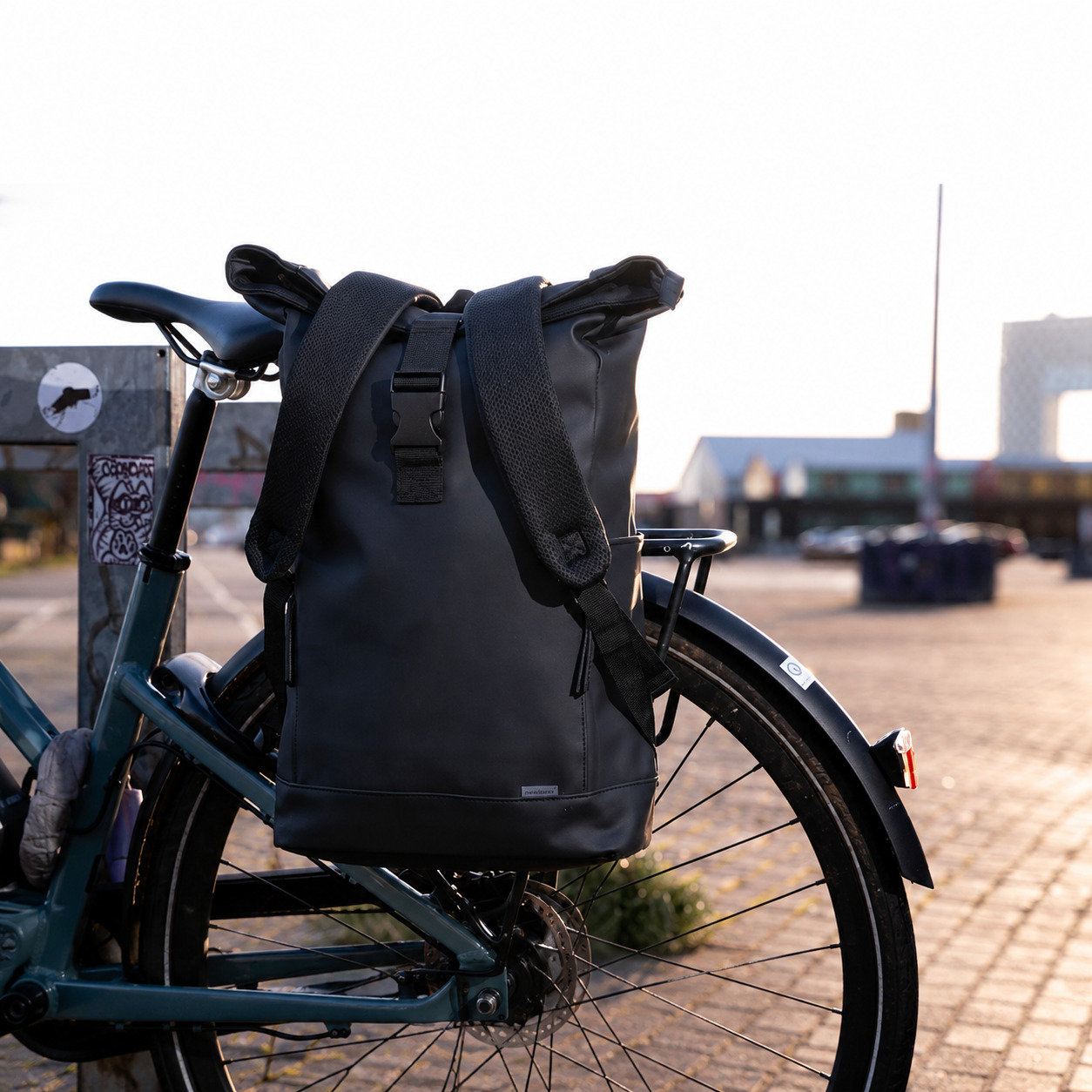 Ein schwarzer Rucksack ist auf dem Gepäckträger eines dunkelgrünen Fahrrads befestigt, im Hintergrund eine Stadt.