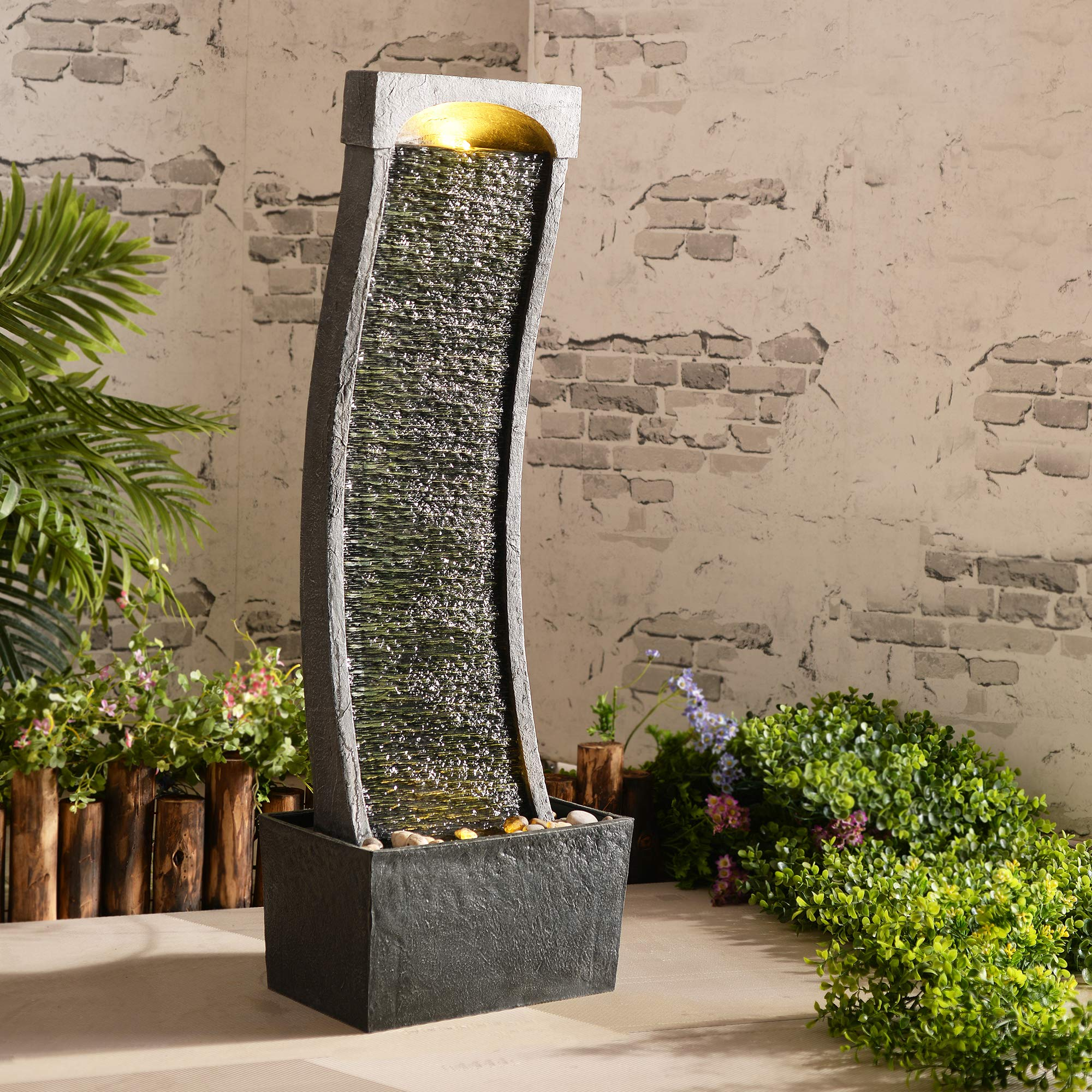Une fontaine d'intérieur grise et courbée avec éclairage se dresse devant un mur de briques, entourée de plantes.