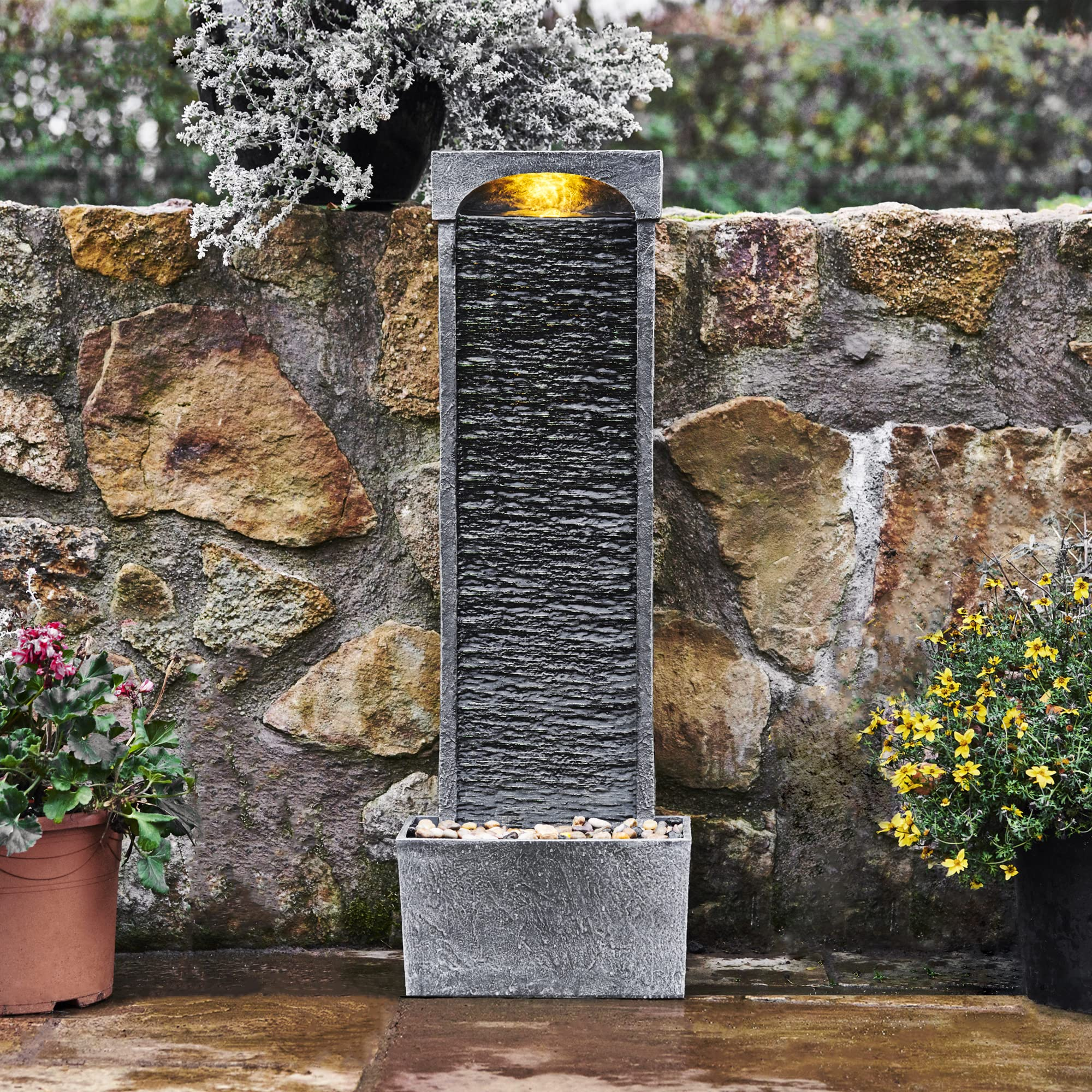 Une fontaine de jardin en pierre éclairée devant un mur en pierre naturelle, encadrée de plantes en pot.