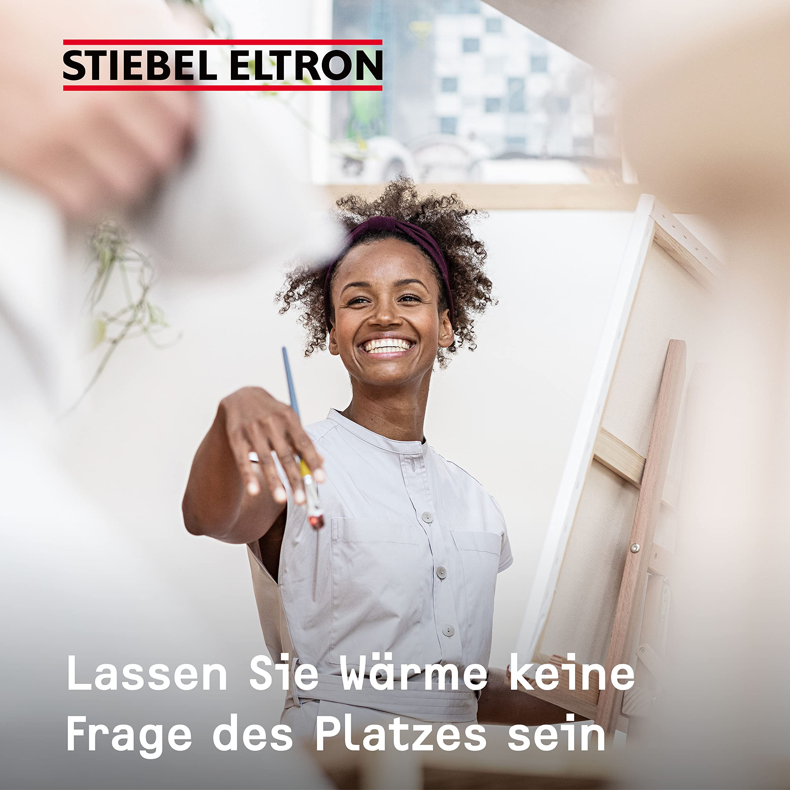 Lächelnte Frau hält einen Pinsel vor einer Leinwand, mit Stiebel Eltron Logo.