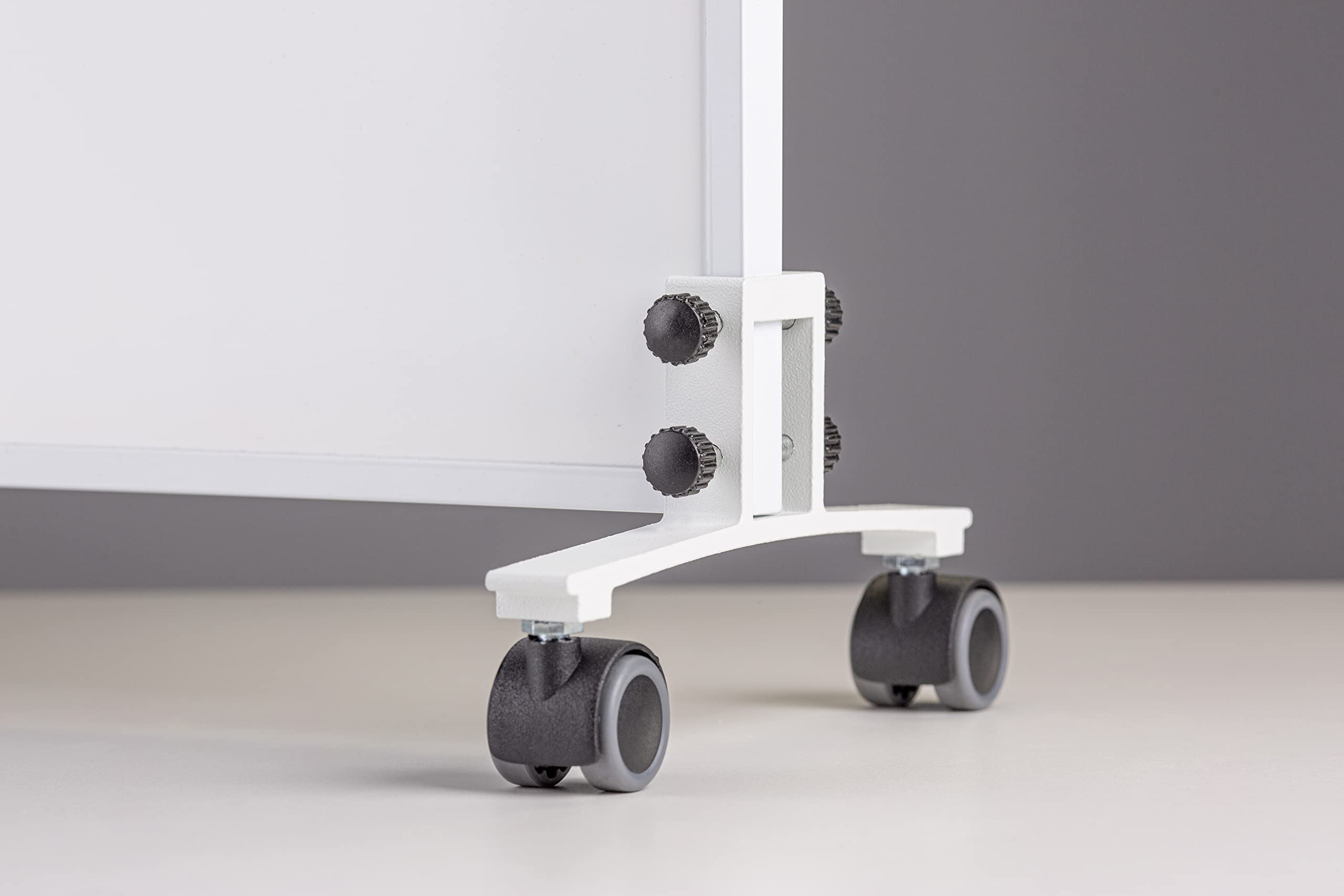 Nahaufnahme eines weißen mobilen Whiteboard-Fußes mit schwarzen Rollen und Einstellknöpfen vor einer grauen Wand.