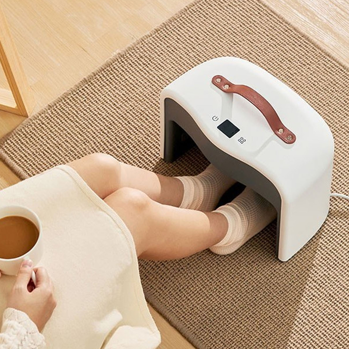 Person sitzt auf einem Teppich, hält eine Kaffeetasse und wärmt die Füße in einem weißen Heizgerät.