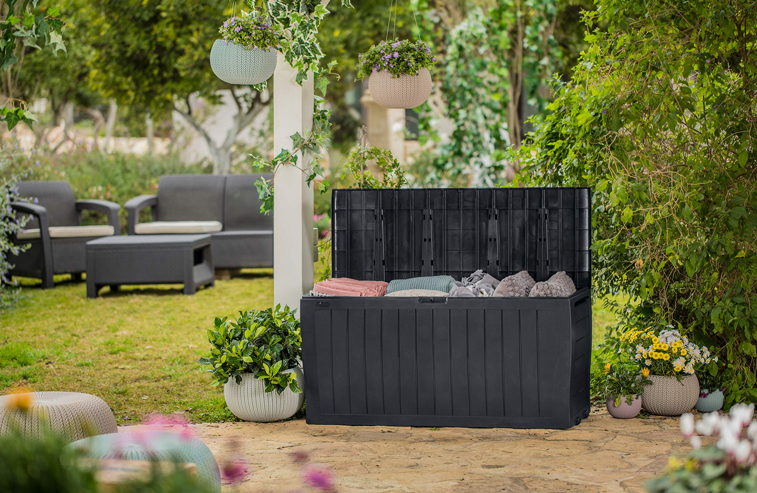Eine offene schwarze Kunststoff-Gartenbox gefüllt mit Kissen steht im Garten vor Pflanzen und Hängekörben.