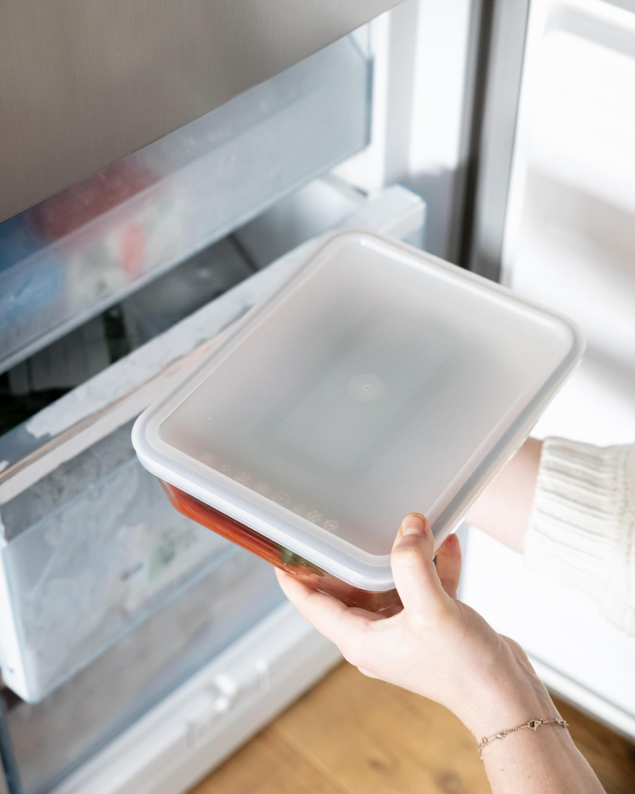 Eine Person hält eine geschlossene Frischhaltebox mit Lebensmitteln vor einem geöffneten Kühlschrank.