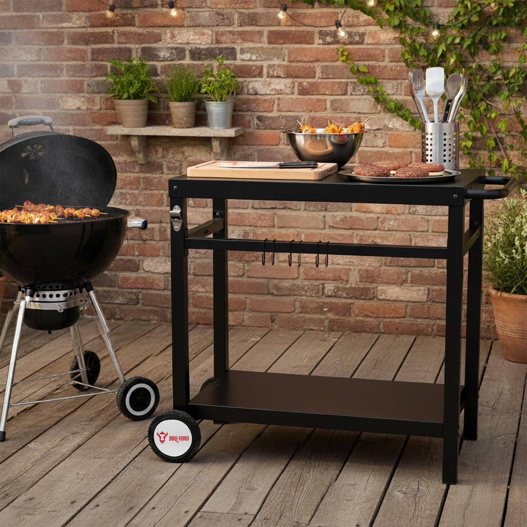 Ein schwarzer Grill und Grillwagen auf Holzdeck vor Backsteinwand mit Grillutensilien.