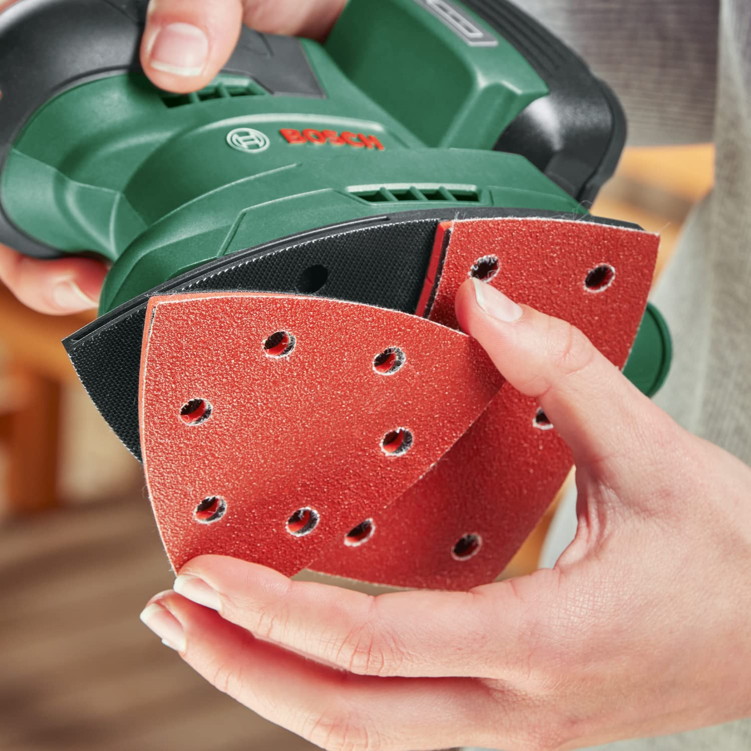 Person wechselt das rote Schleifpapier an einem grünen Bosch-Multischleifer.