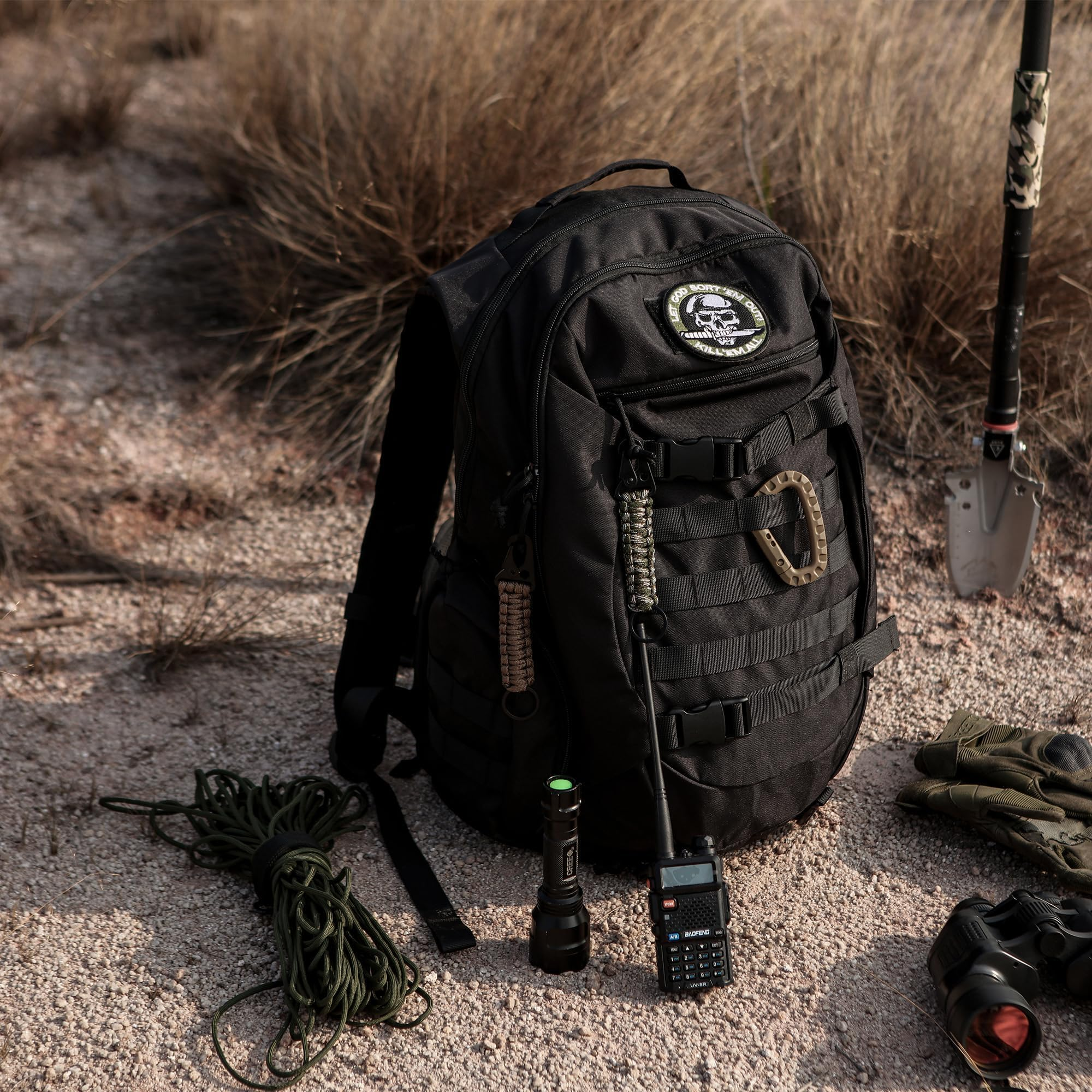 Ein schwarzer Rucksack mit Abzeichen, Taschenlampe, Funkgerät und Seil auf felsigem Boden vor Gras.