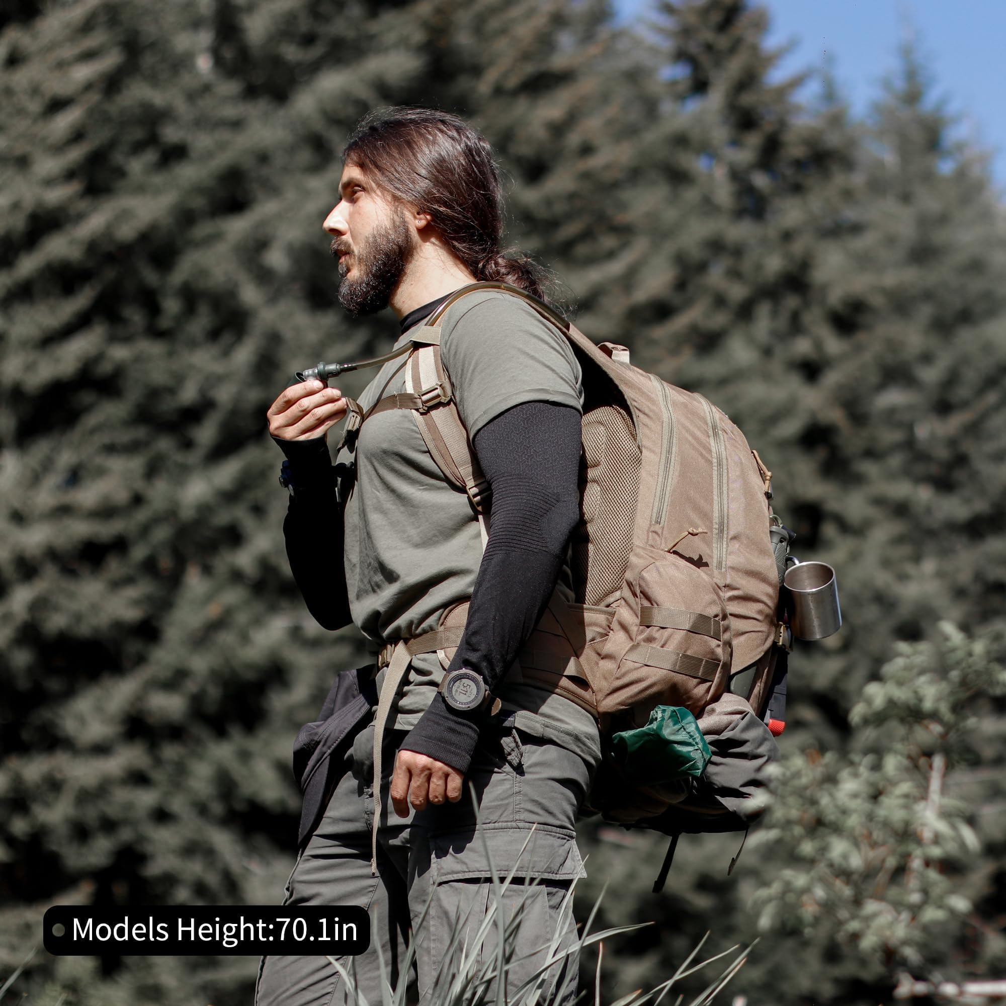 Ein langhaariger Mann mit Bart und großem Rucksack steht im Wald vor grünen Bäumen.