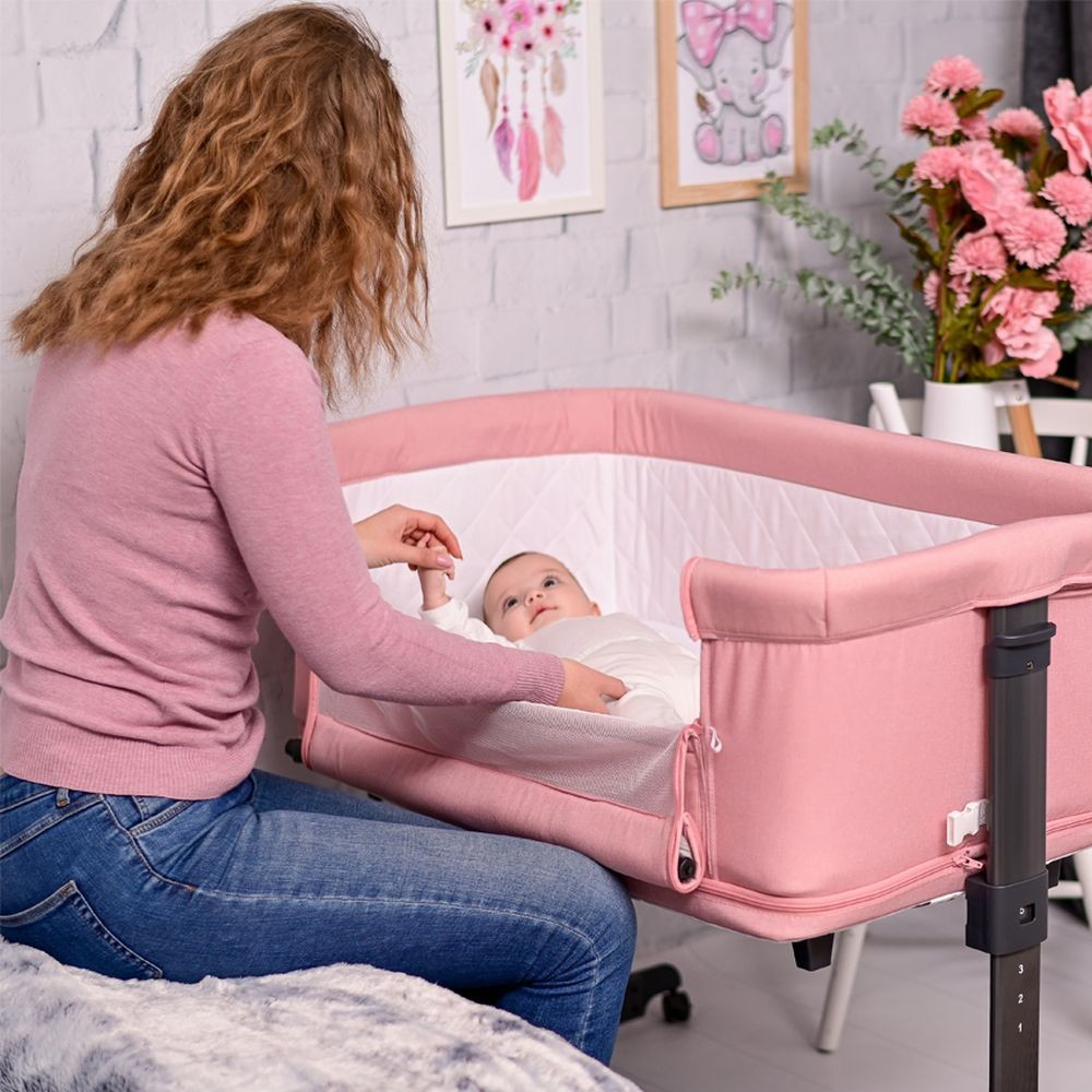 Eine Mutter im rosa Pullover sitzt neben einer rosa Wiege und hält die Hand ihres Babys in einem hellen Kinderzimmer.