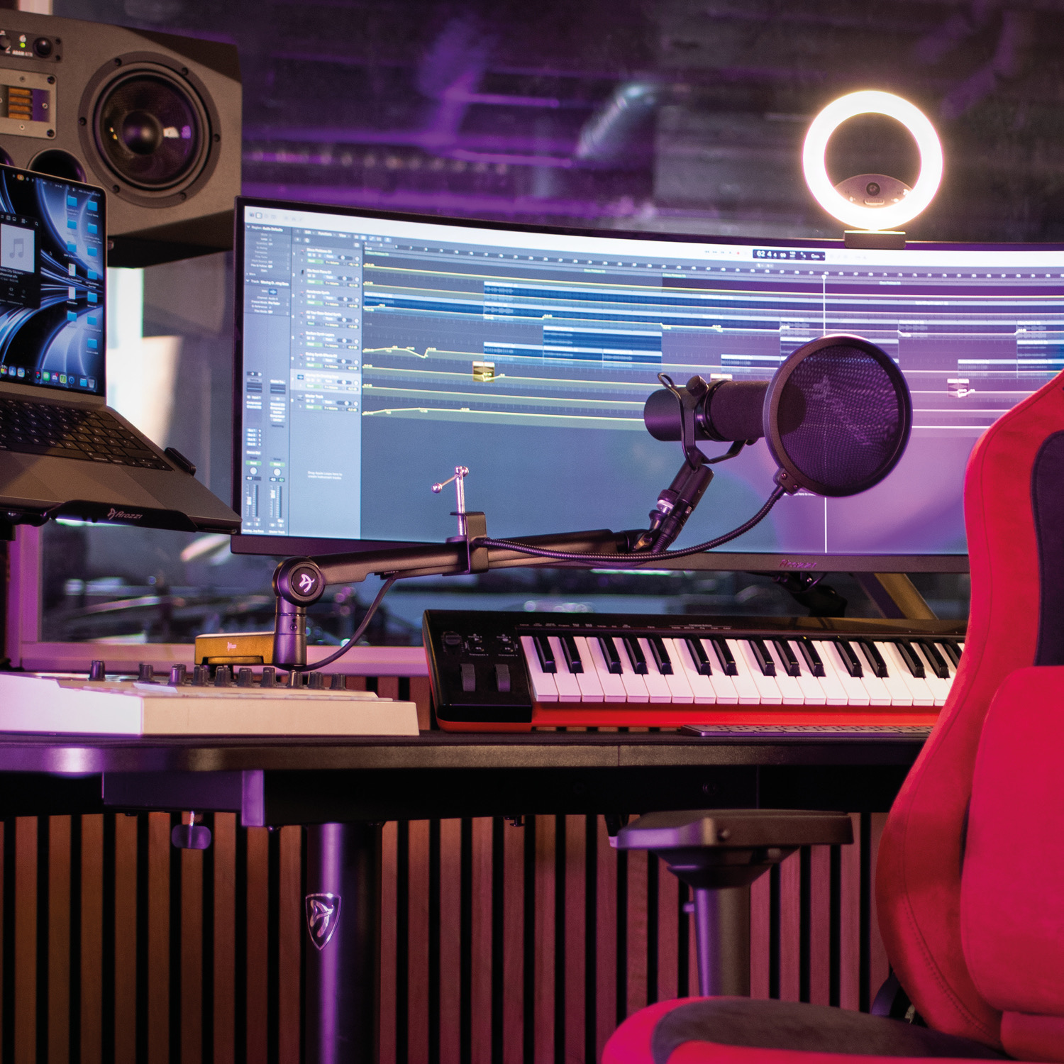 Ein Musikstudio mit gebogenem Bildschirm, Laptop, Mikrofon, MIDI-Keyboard, Lautsprecher und rotem Stuhl.