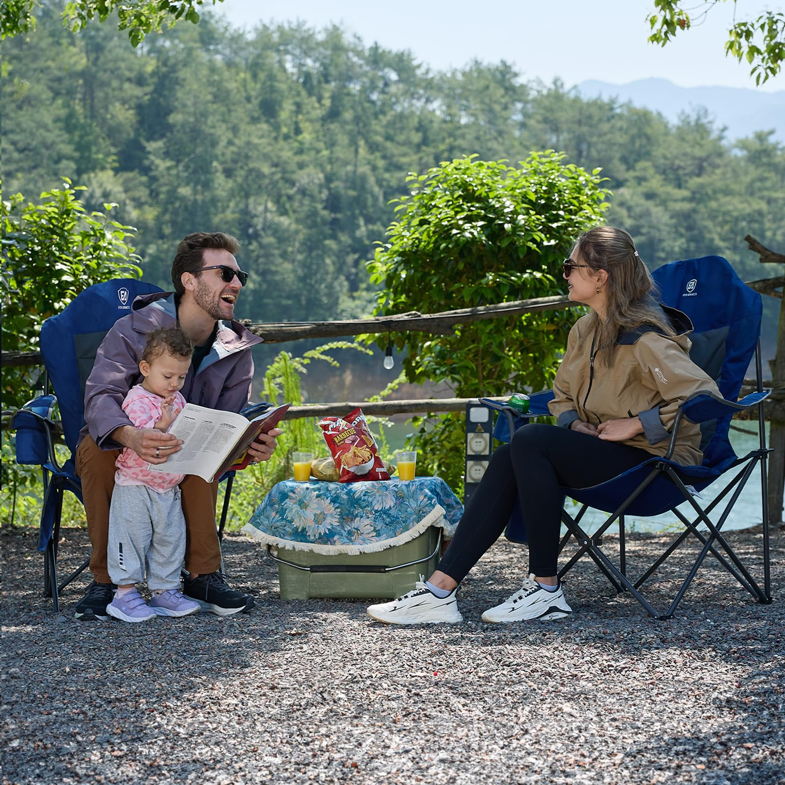 Una famiglia in campeggio vicino a un lago, seduta su sedie blu, legge un libro e mangia patatine tra i monti.