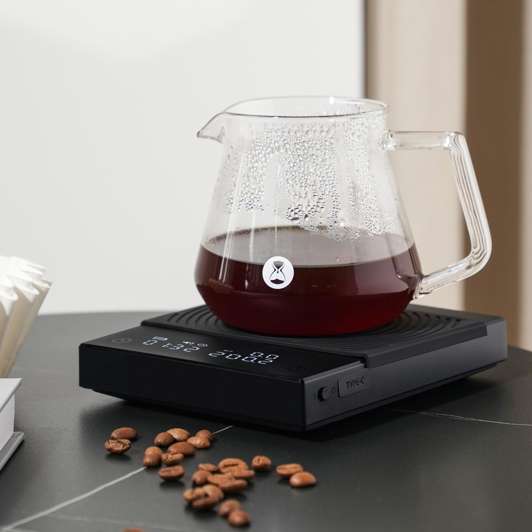 Eine Glaskanne mit gebrühtem Kaffee steht auf einer schwarzen Digitalwaage, umgeben von Kaffeebohnen auf einem Tisch.