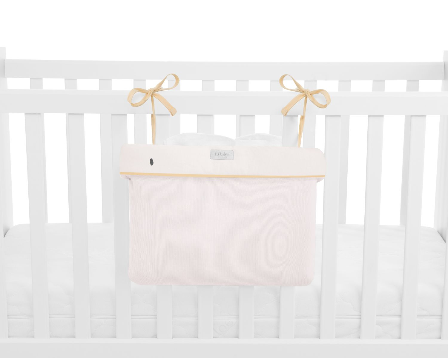 Ein hellrosa Baby-Stofforganizer hängt an den weißen Gittern eines Babybetts, befestigt mit beigen Schleifen.