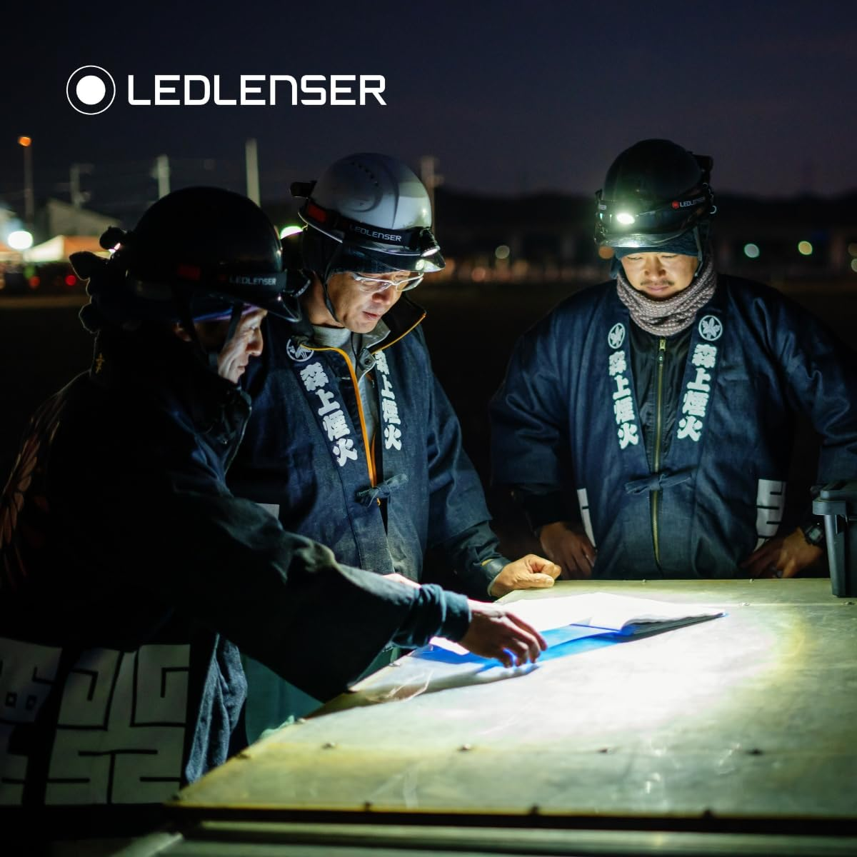 Drei Arbeiter mit Stirnlampen schauen nachts bei hellem Licht auf Dokumente.