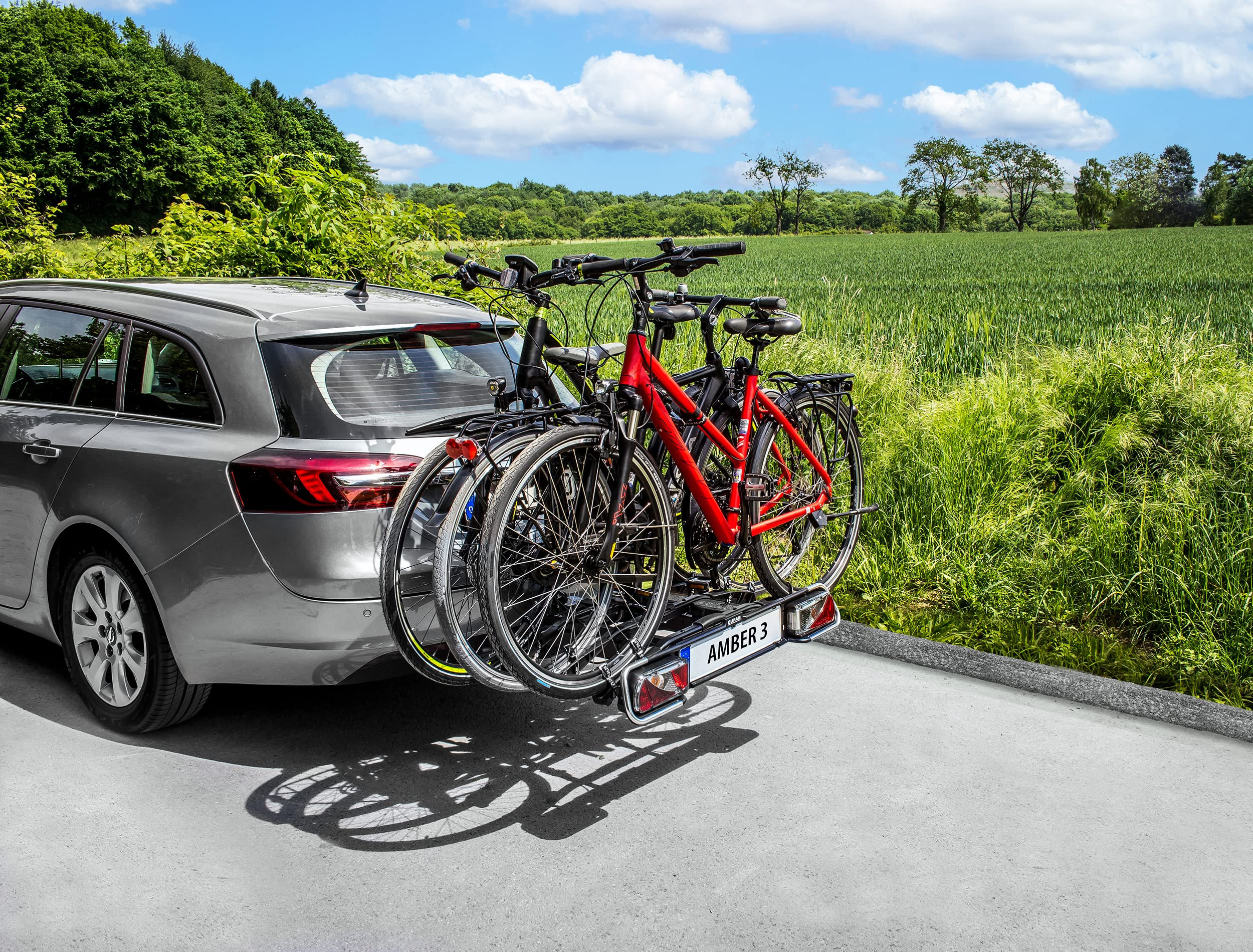 Un'auto grigia con un portabici posteriore trasporta tre biciclette su una strada asfaltata davanti a un paesaggio verde.