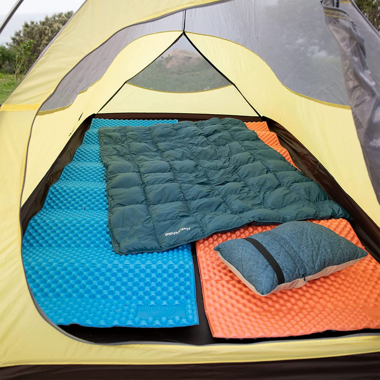 Interno di una tenda gialla con materassini blu e arancioni, una coperta verde e un cuscino.