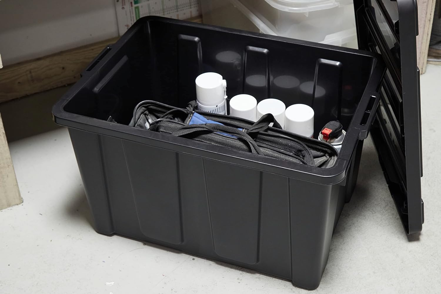 Una scatola di plastica nera contenente una borsa portautensili e diverse bombolette spray, su un pavimento chiaro.