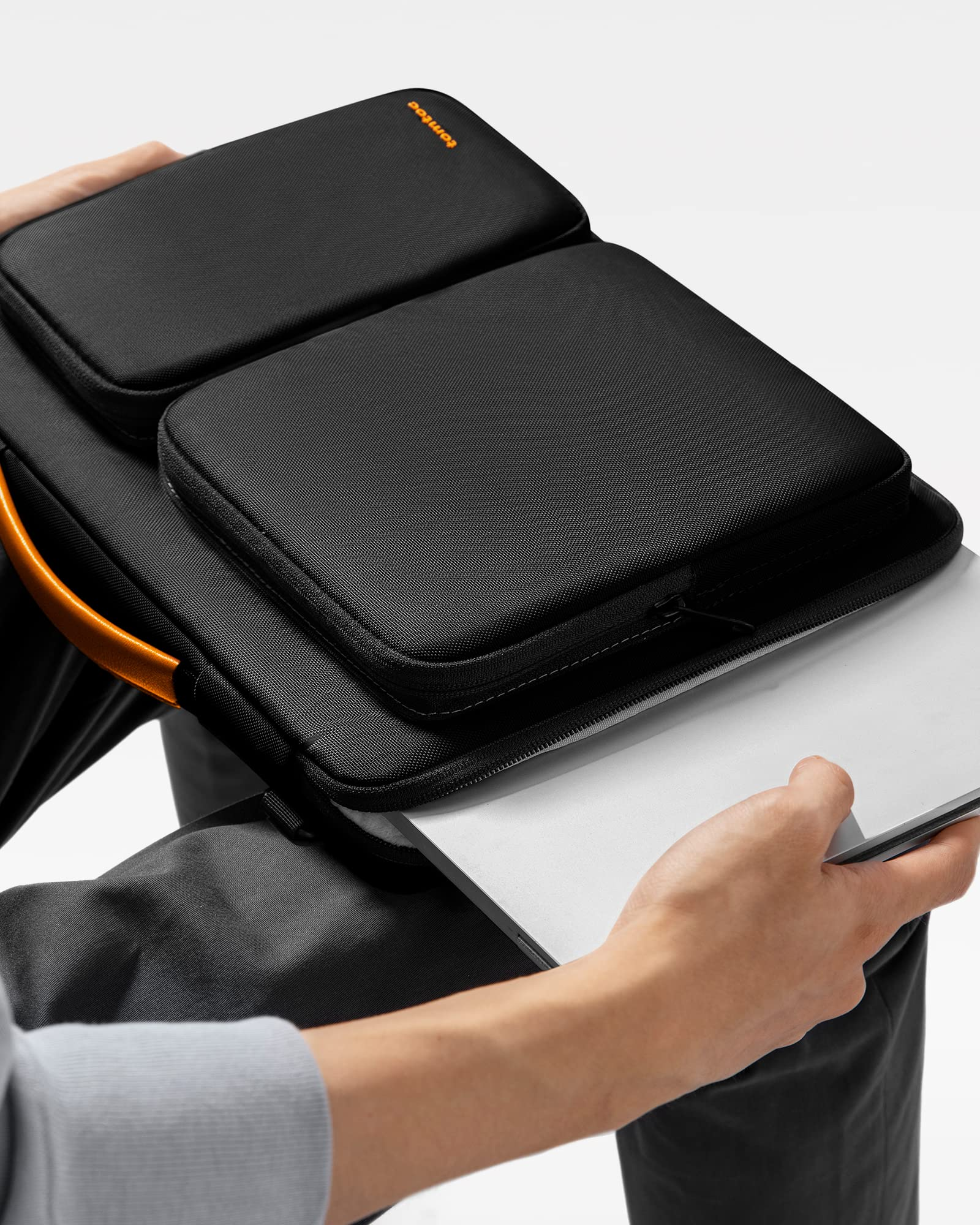 Eine Person legt einen silbernen Laptop in eine schwarze Tasche mit zwei Außentaschen und orangem Griff.