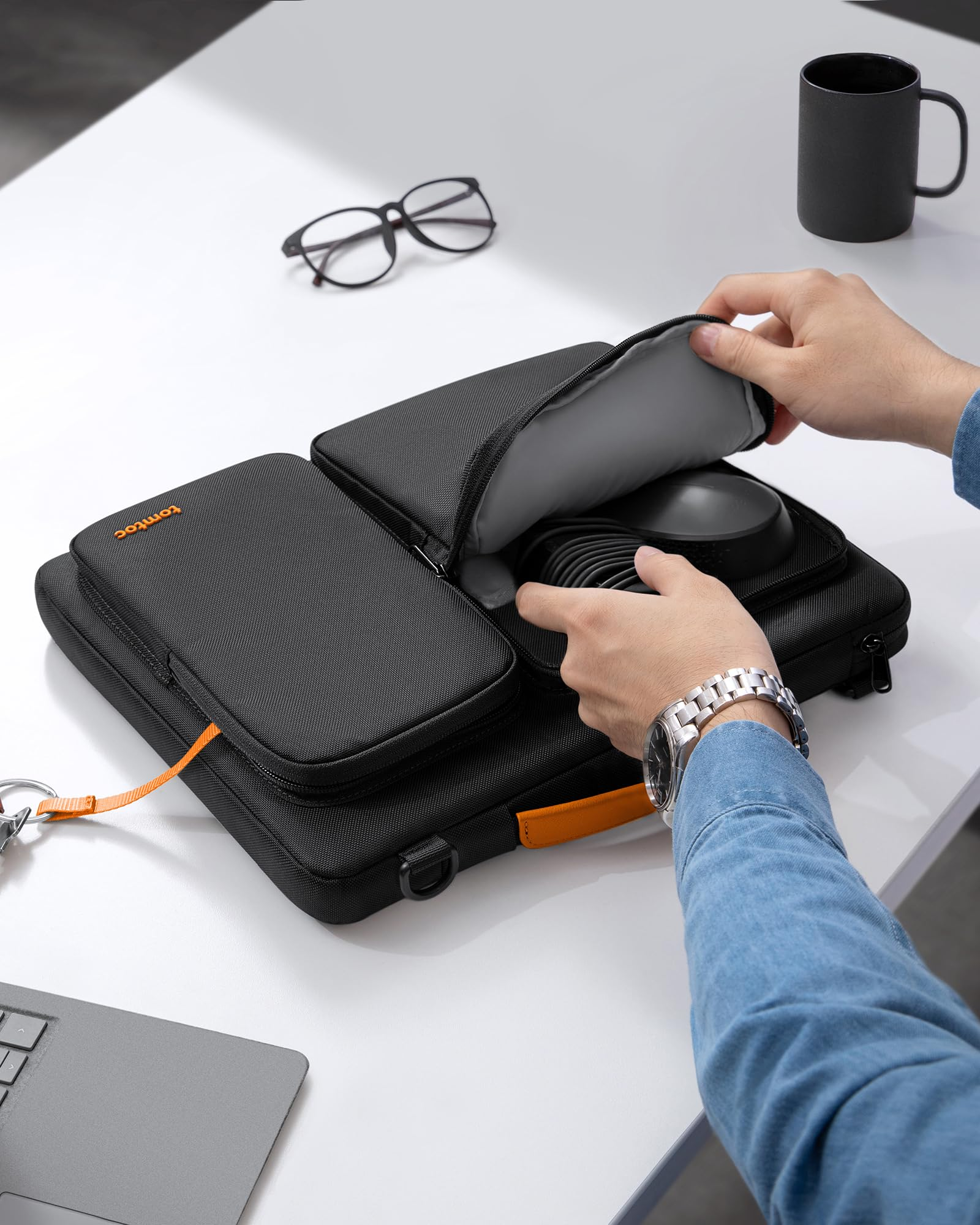 Person öffnet eine schwarze Laptoptasche mit Zubehör auf einem weißen Tisch.