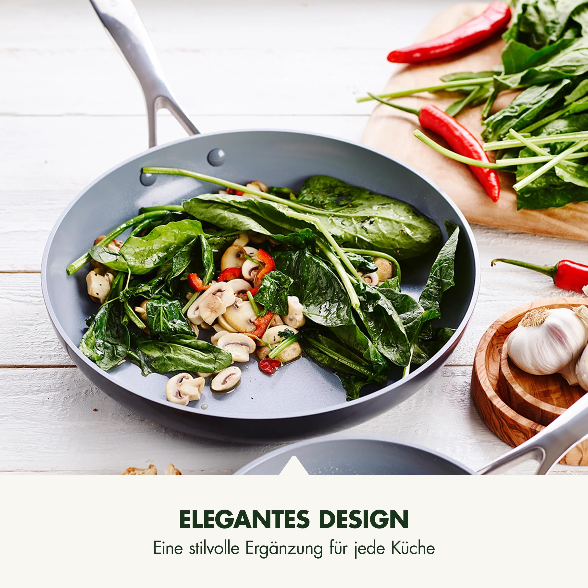 Une poêle grise avec des épinards et des champignons frais sur une table en bois, à côté de piments et d'ail.