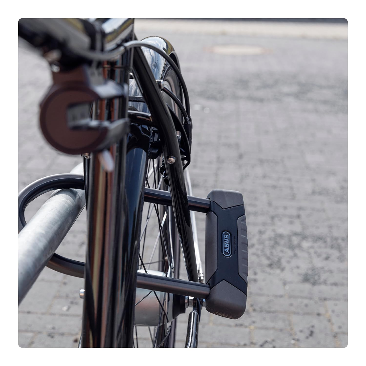 Un vélo noir attaché à un support en métal avec un antivol en U Abus sur une rue pavée.