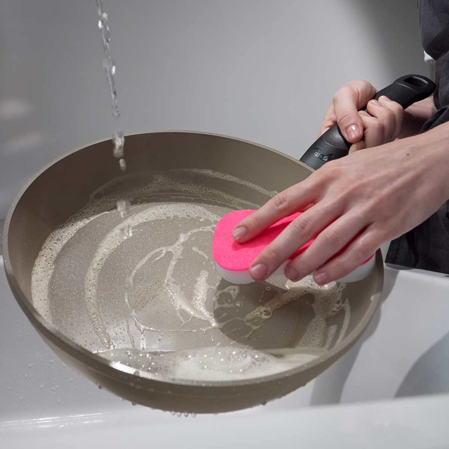 Une personne lave une poêle marron avec de la mousse de savon et une éponge rose sous l'eau courante.