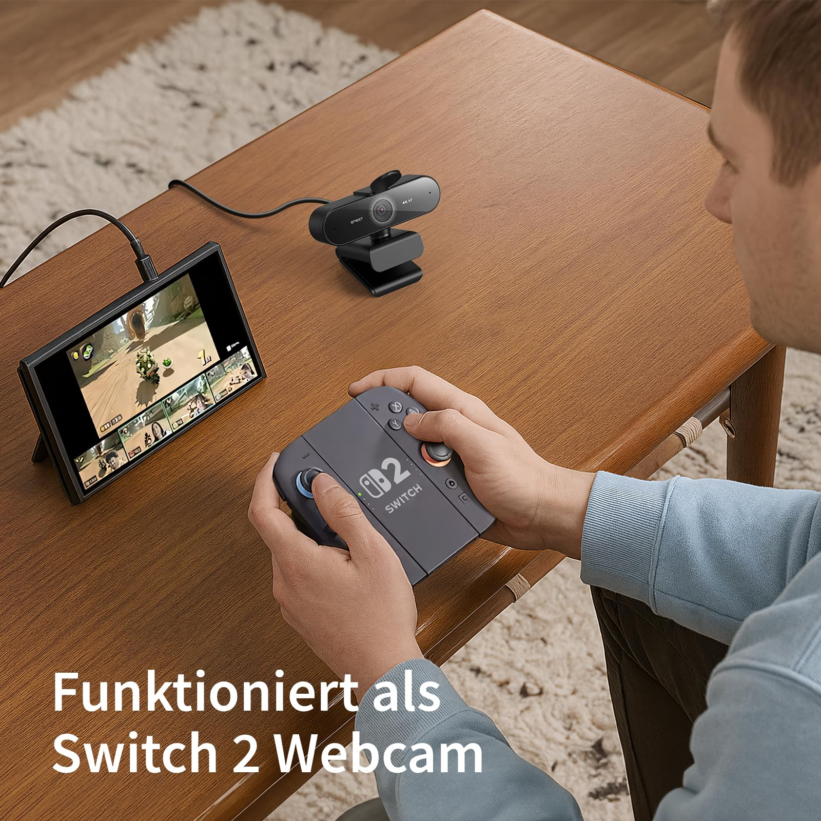 Un uomo siede a un tavolo di legno tenendo un controller grigio, con uno schermo e una webcam sulla sinistra.