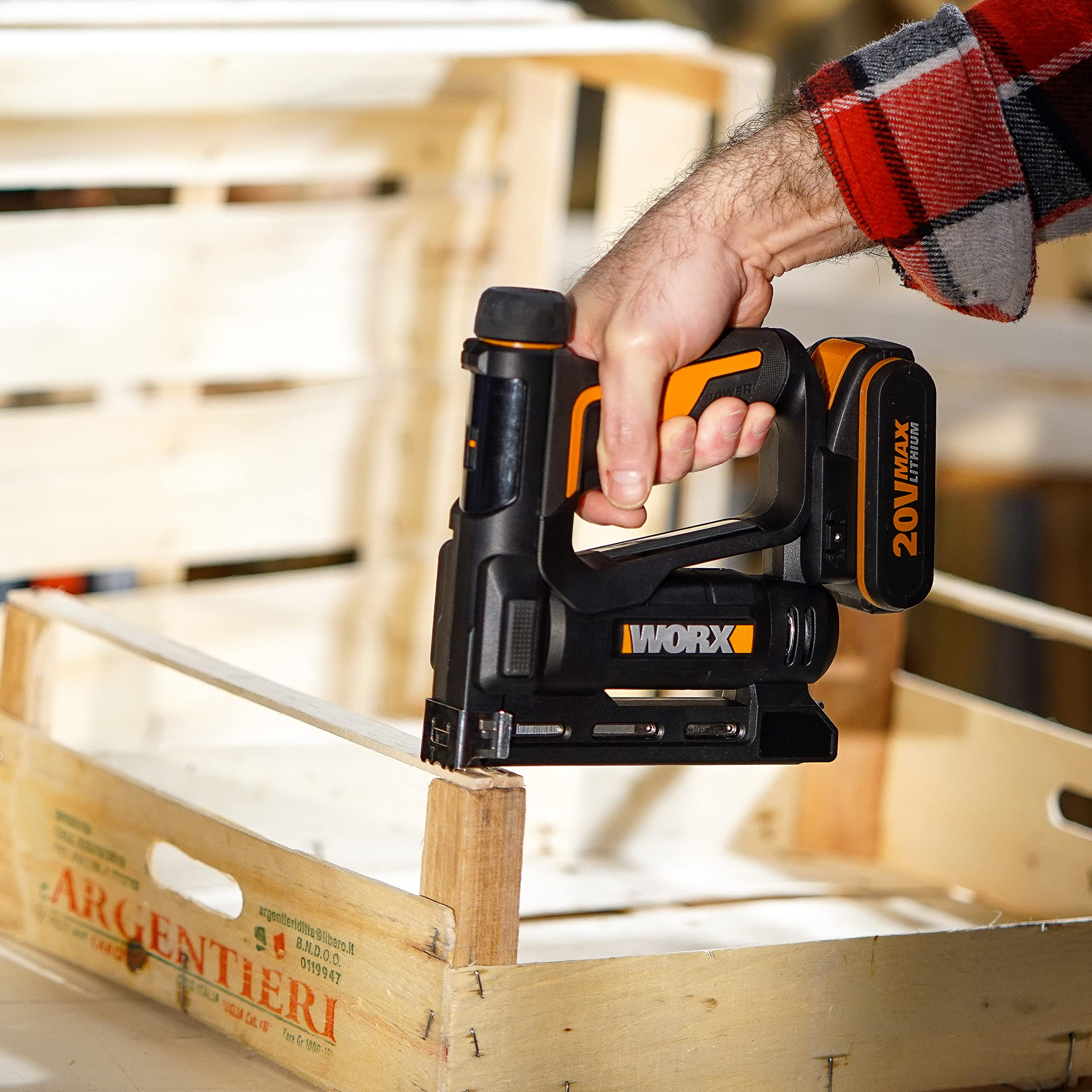 Eine Hand im karierten Hemd hält einen schwarzen und orangen Akku-Tacker über einer Holzkiste.