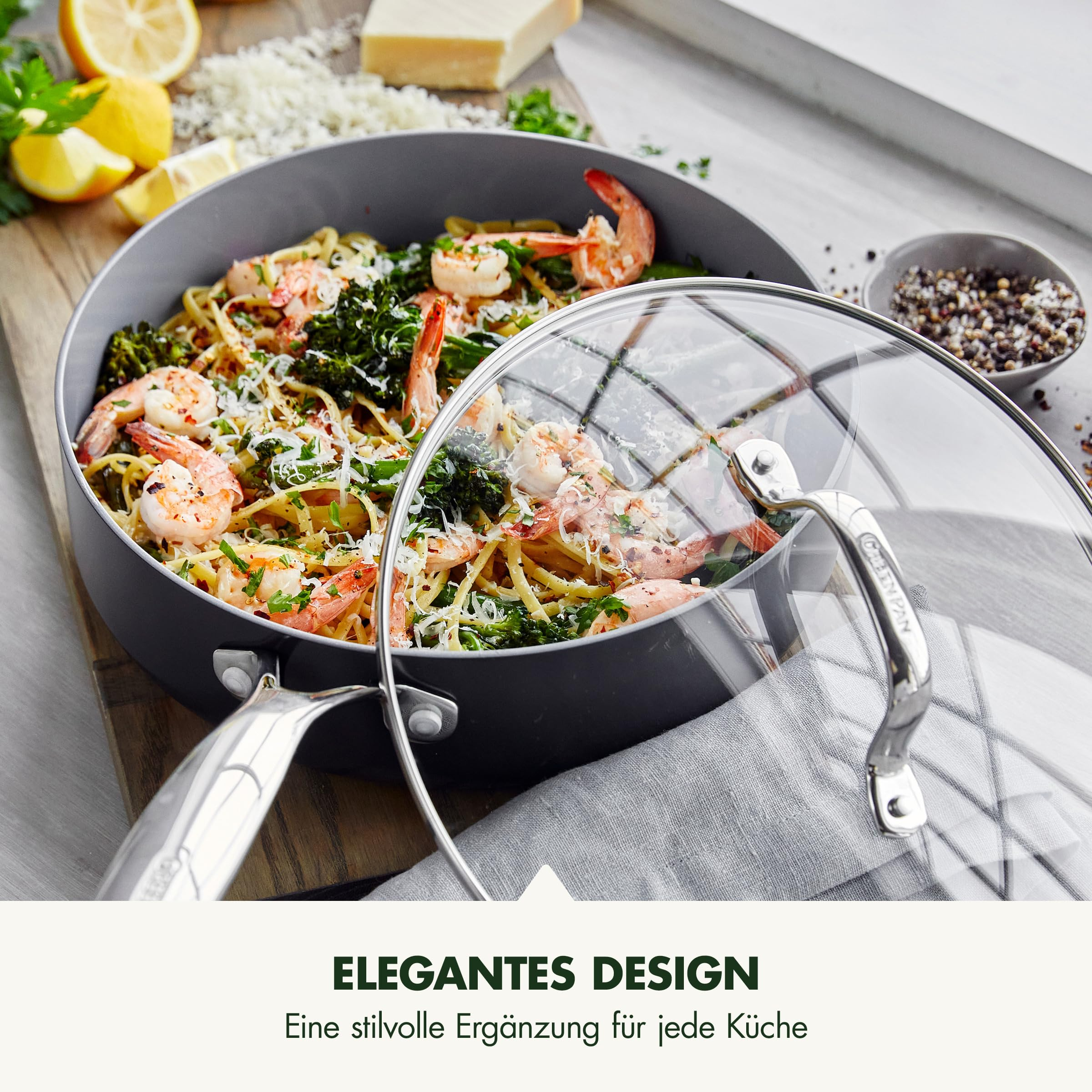 Une poêle grise avec crevettes, pâtes et légumes, couvercle en verre et citrons sur planche.