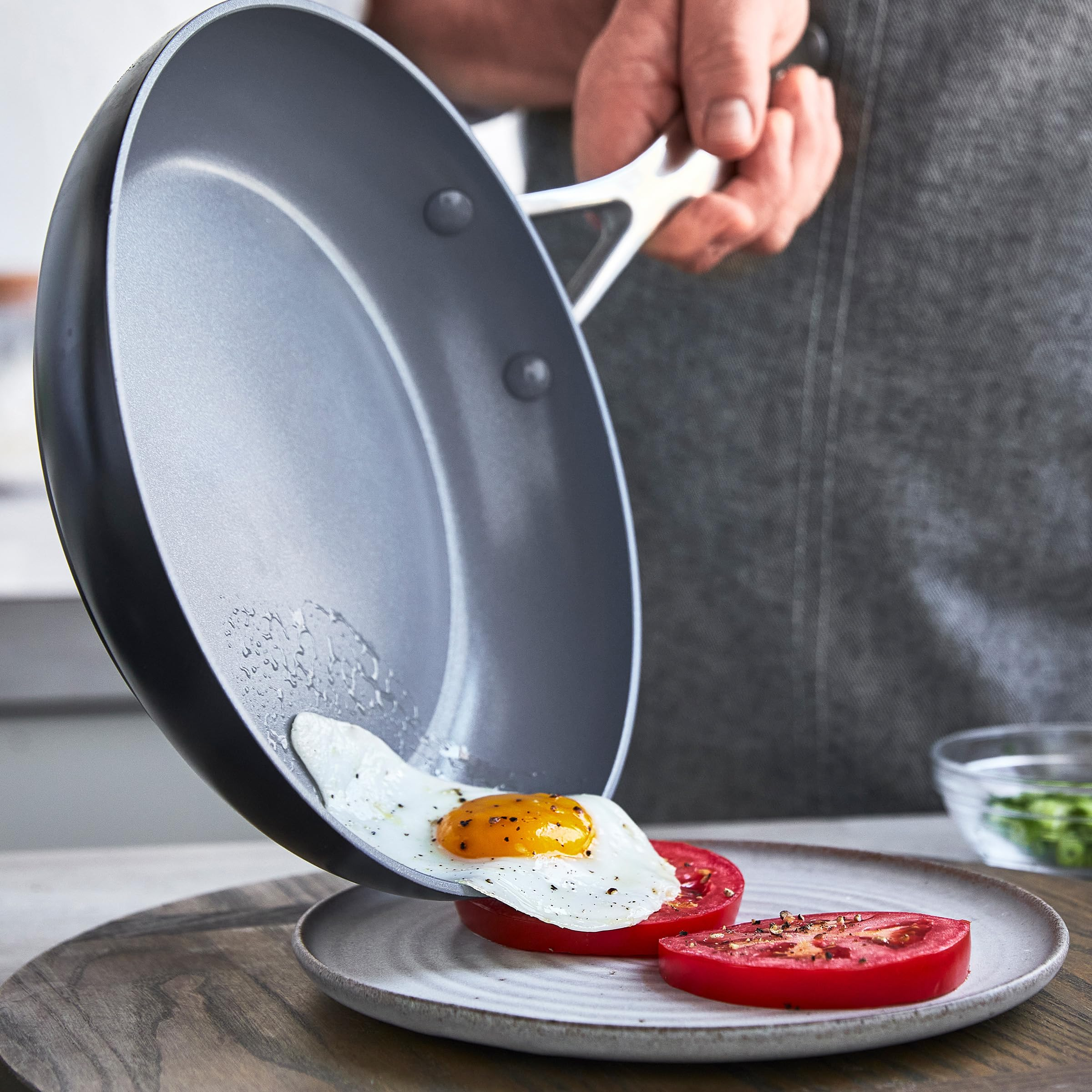 Un chef fait glisser un œuf au plat d'une poêle grise sur des tomates en tranches dans une assiette.