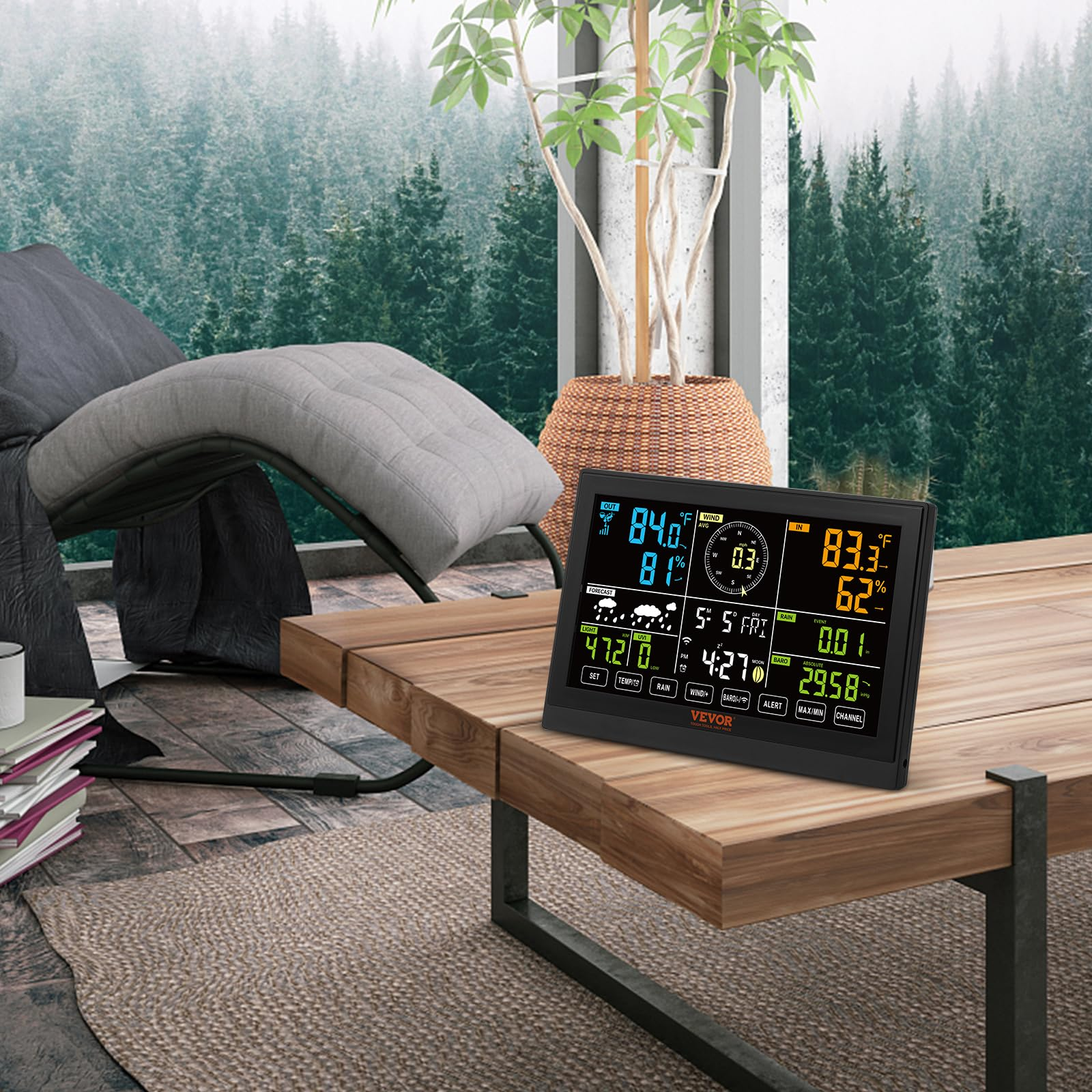 Station météo sur une table en bois devant un paysage forestier et un fauteuil.