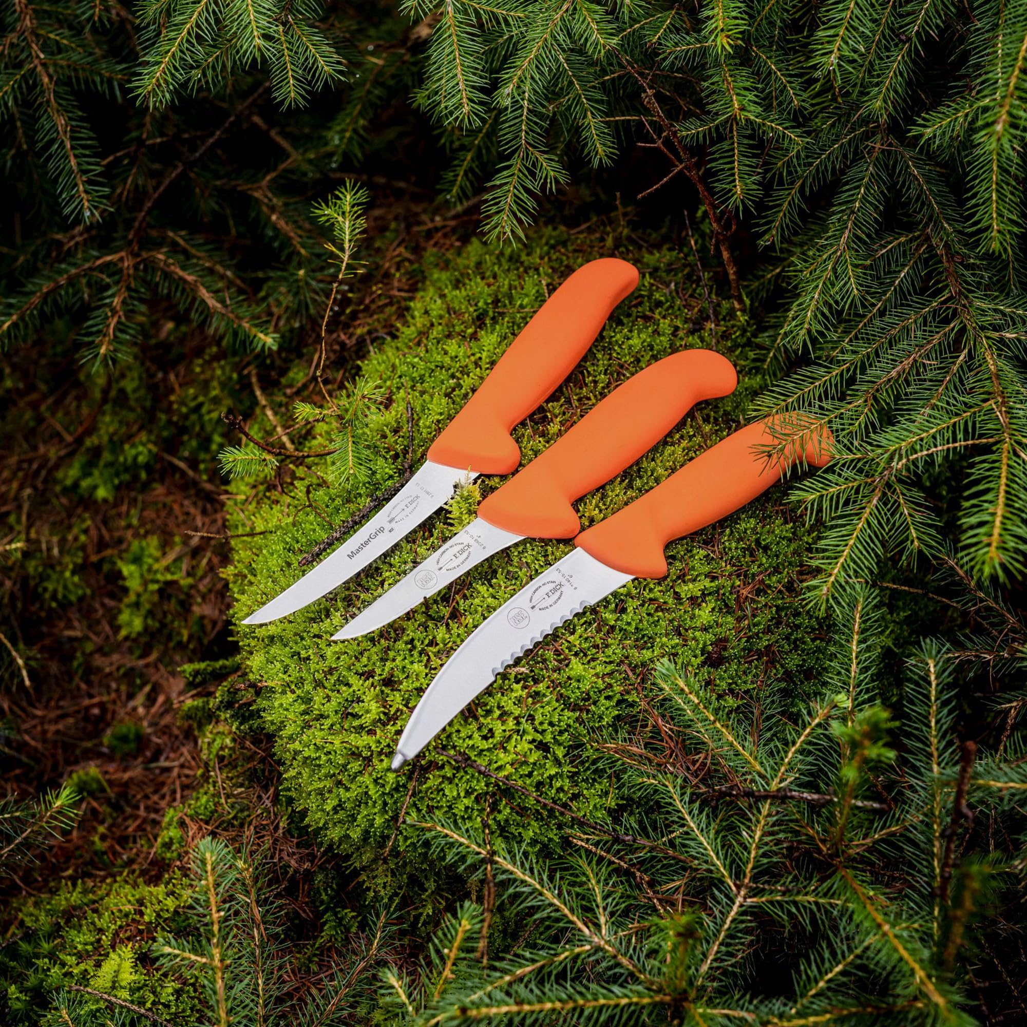 Drei orange bemaschte Messer liegen nebeneinander auf Moos, umgeben von grünen Nadelbaumzweigen.