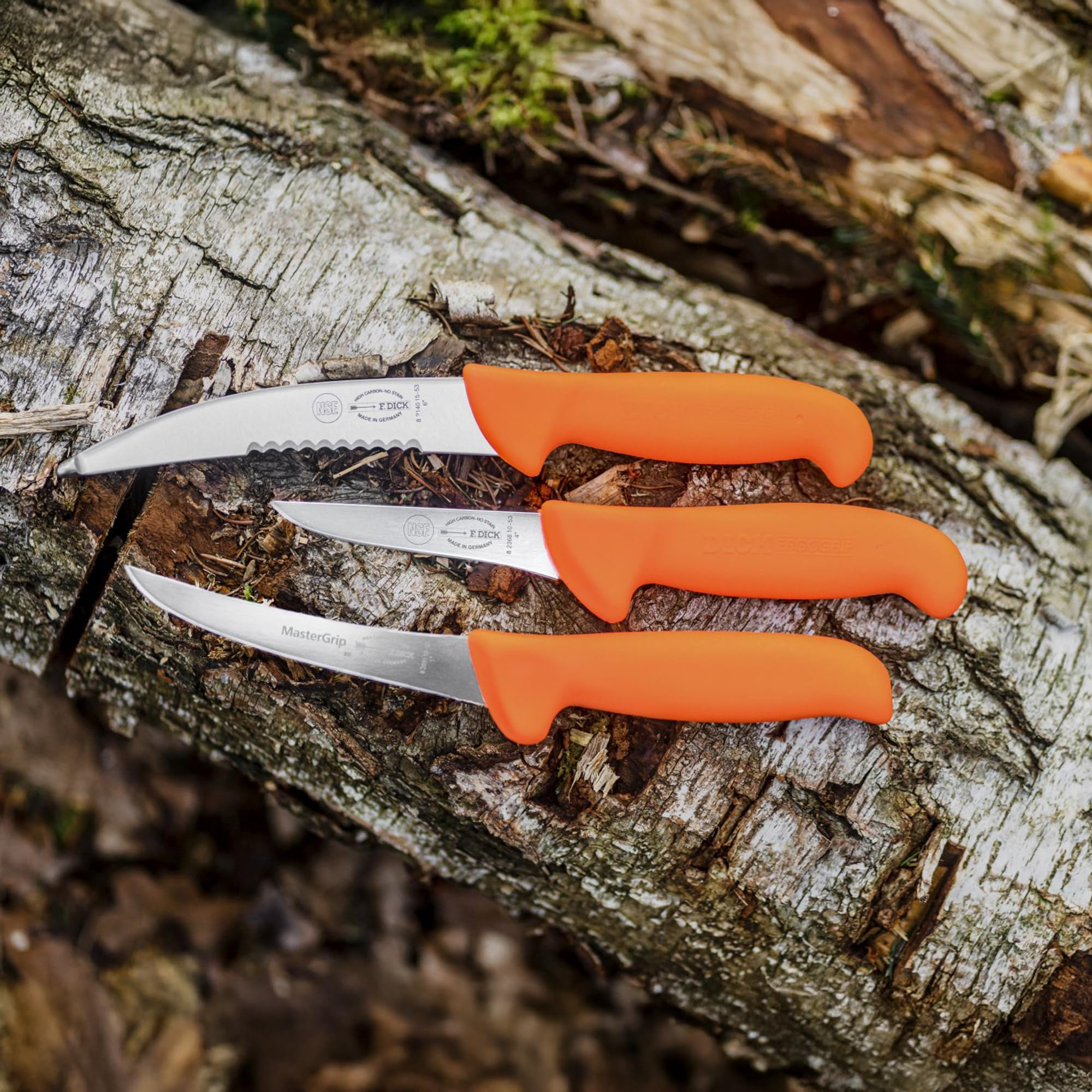 Drei Jagdmesser mit leuchtend orangefarbenen Griffen liegen auf einer rauen Baumrinde.
