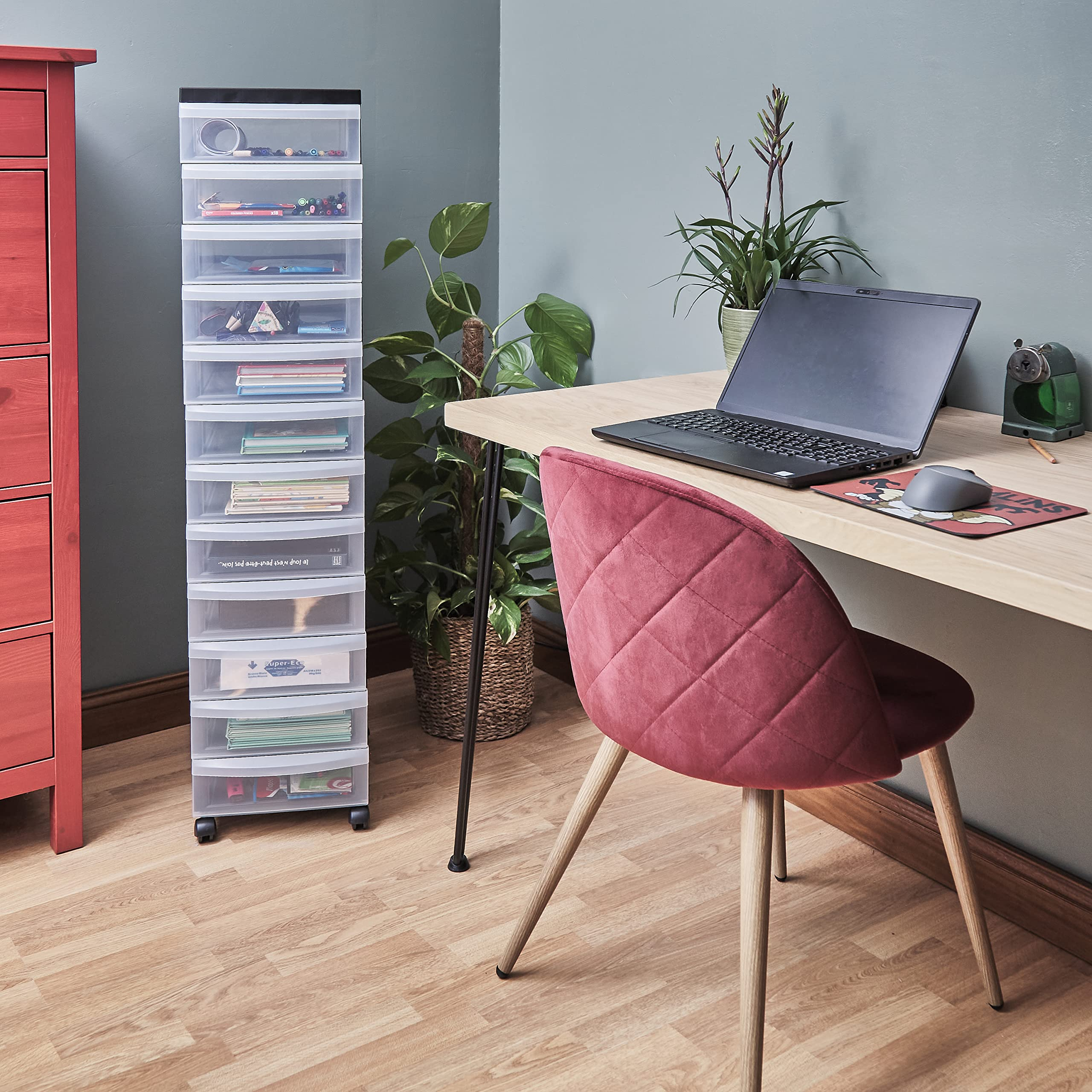 Ein Heimbüro mit Holztisch, rotem Stuhl, Laptop, Pflanzen und einem hohen Rollschubladenschrank in einer Ecke.