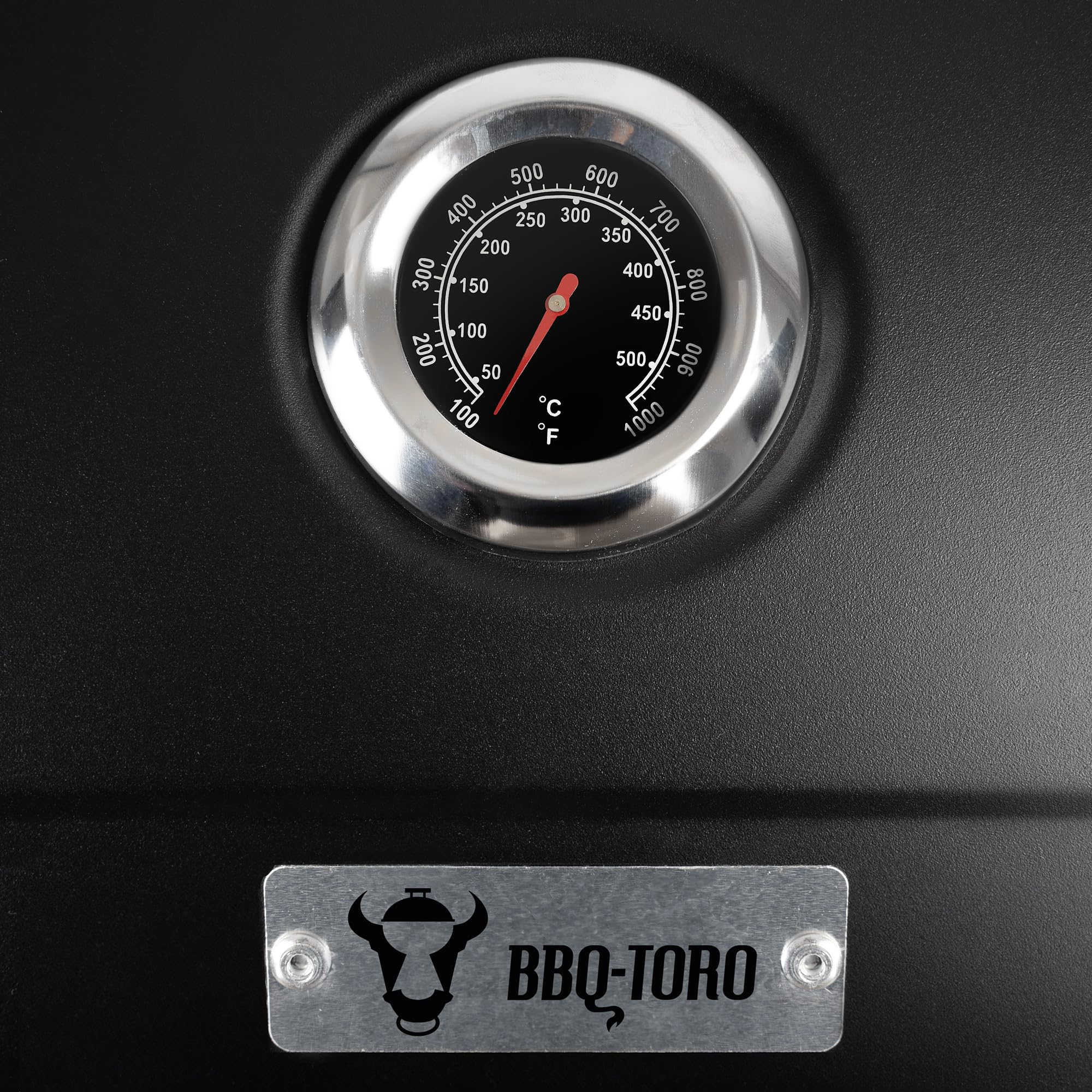 Un thermomètre de grill rond argenté sur fond noir mat, avec une plaque avec le logo BBQ-Toro en dessous.