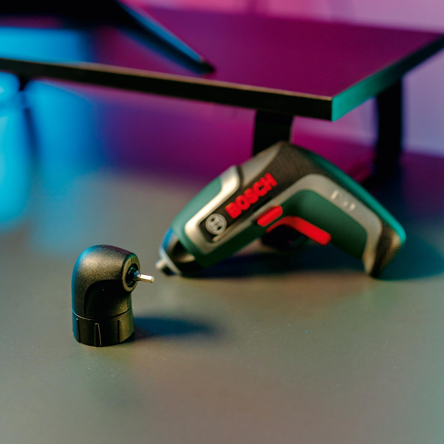 Un avvitatore a batteria Bosch e un accessorio angolare si trovano su una scrivania scura sotto una luce blu e viola.