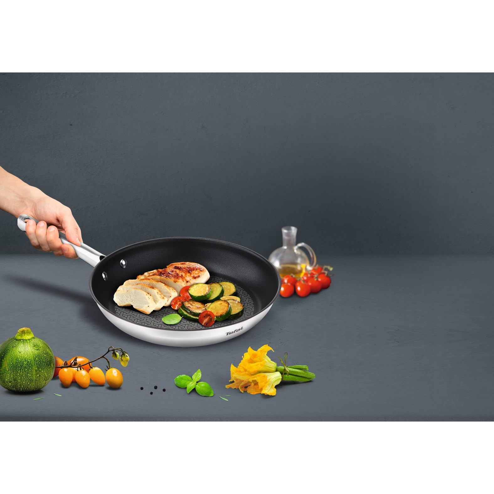 Mano sosteniendo una sartén con pollo y calabacín. Verduras frescas sobre la encimera gris.