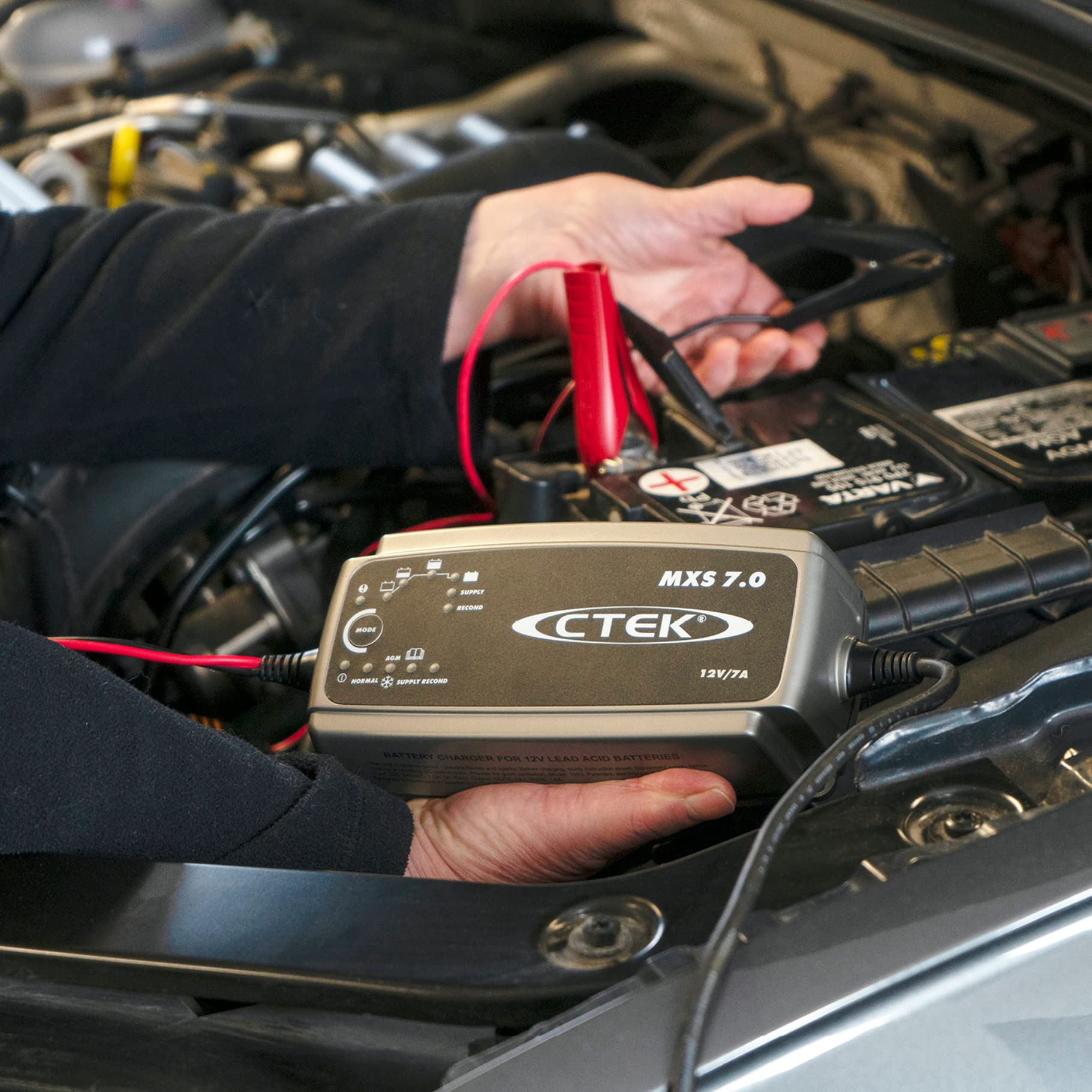 Una persona tiene in mano un caricabatterie CTEK vicino al motore di un'auto e alla batteria con i cavi.