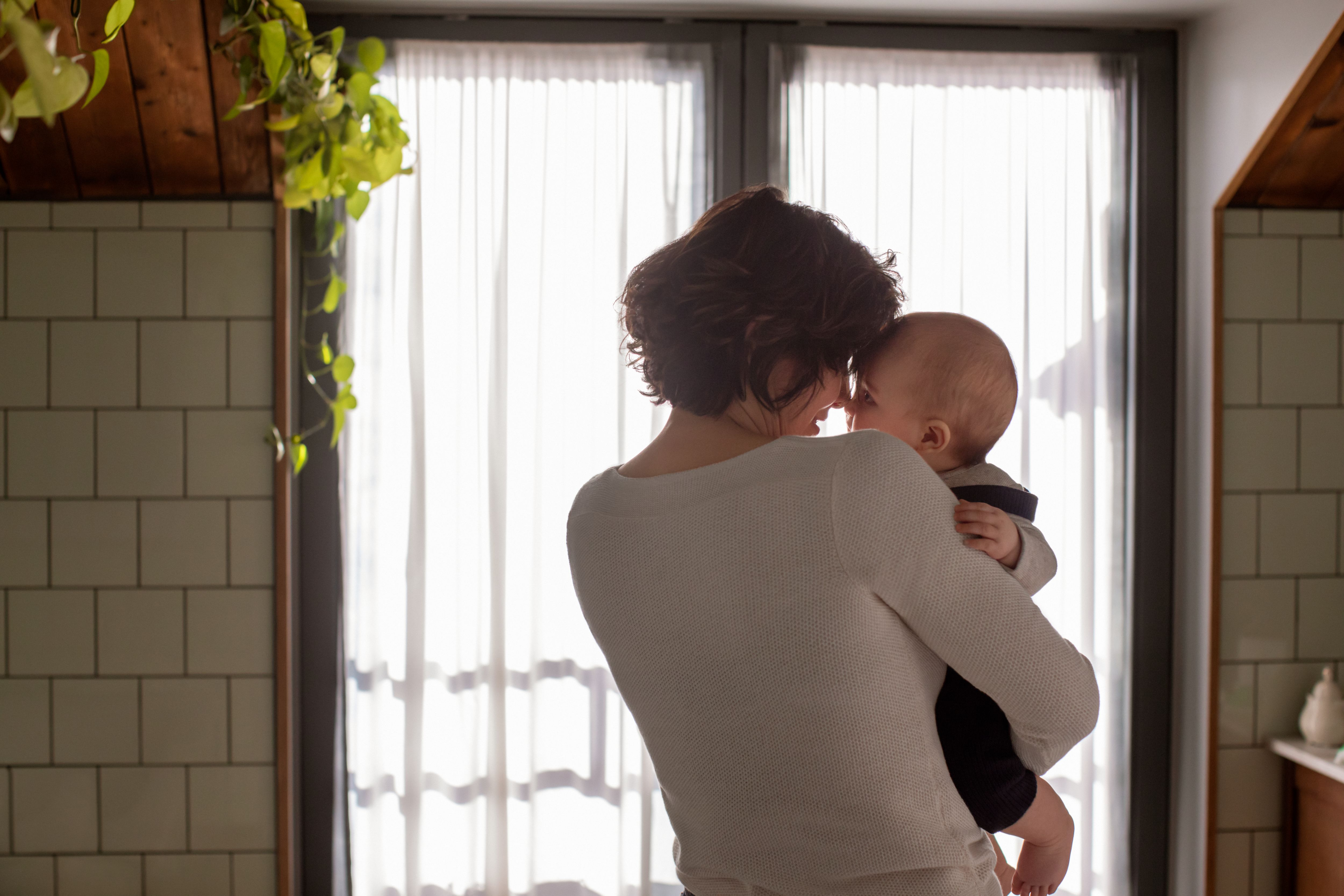 Una madre con un maglione bianco tiene in braccio il suo bambino vicino a una finestra con tende trasparenti, condividendo un momento tenero.