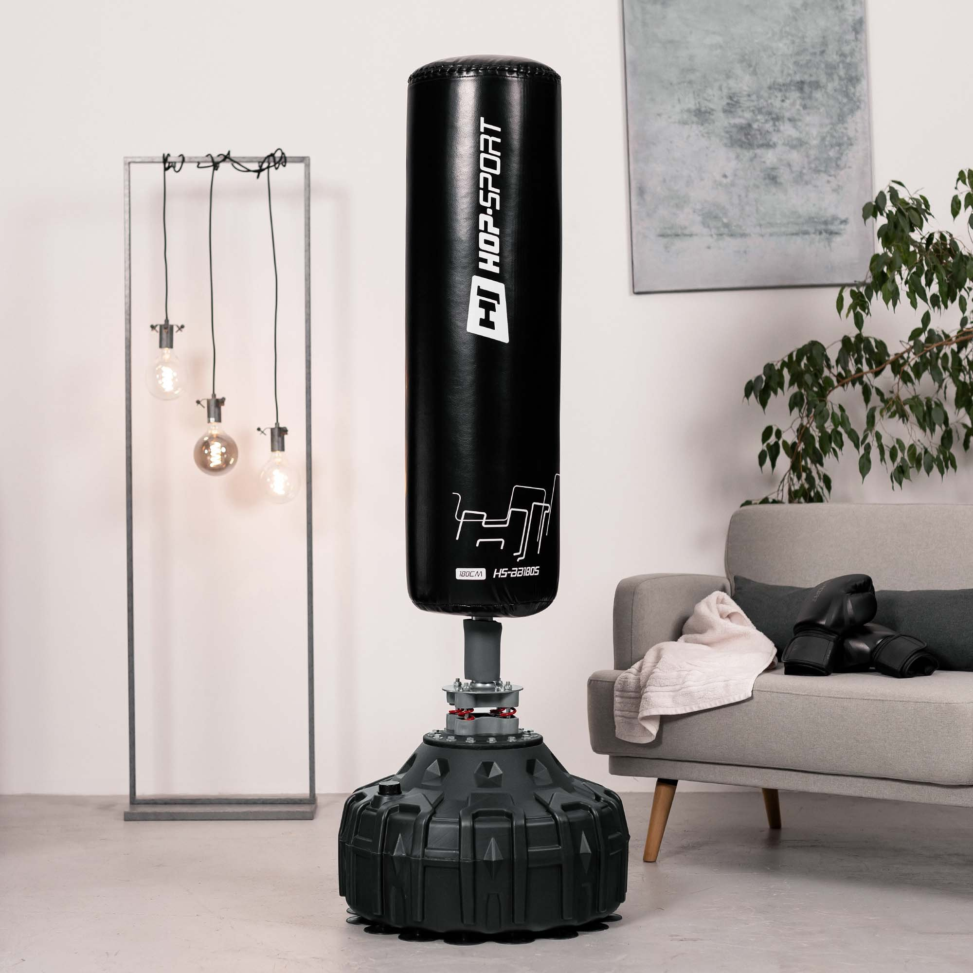 Ein schwarzer Stand-Boxsack HOP-SPORT steht in einem hellen Wohnzimmer mit grauen Möbeln, Pflanzen und Lampen.