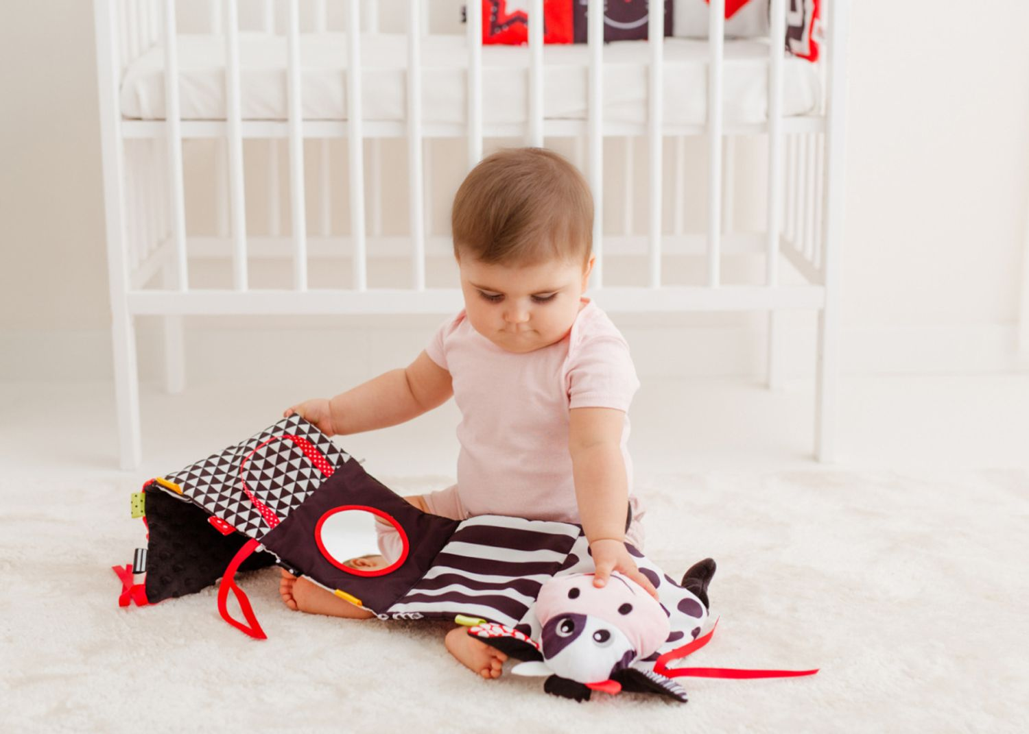 Ein Baby in einem rosa Romper sitzt auf einem weißen Teppich und spielt vor einem Gitterbett mit einem kontrastreichen Spielzeug.